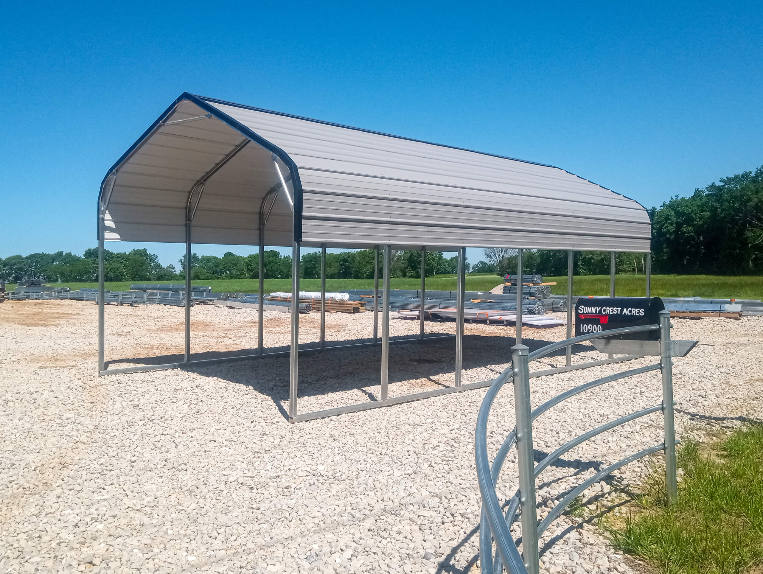 Sunny Crest Acres Versailles MO A metal carport structure stands on a gravel lot under a clear blue sky, with a sign reading "Sunny Crest Acres 10960" visible in the foreground.
