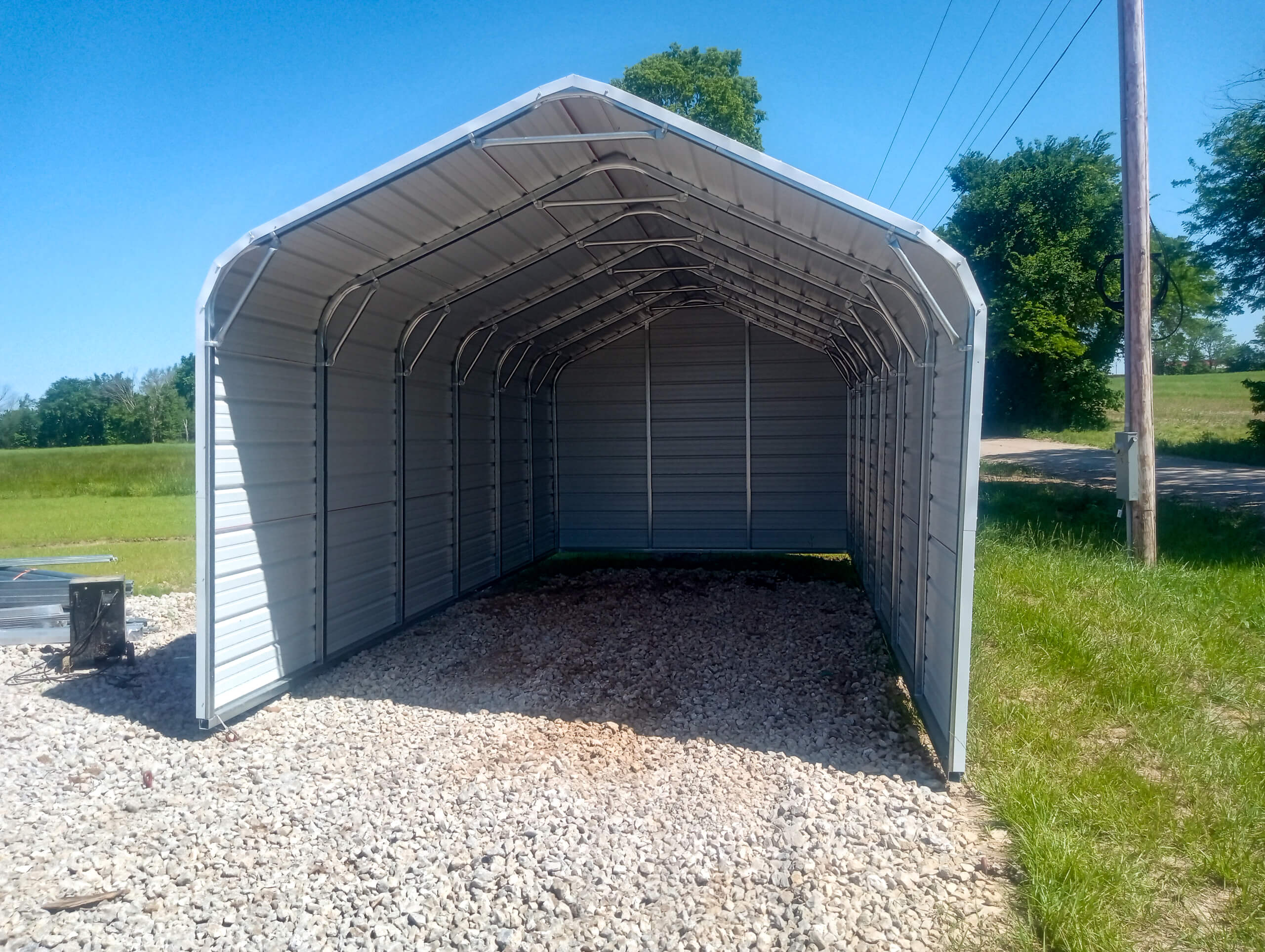 6.1.26 Sunny Crest Acres Gallery 2-3 A metal carport structure with an open front stands on a gravel surface under a clear blue sky, surrounded by grass and trees.