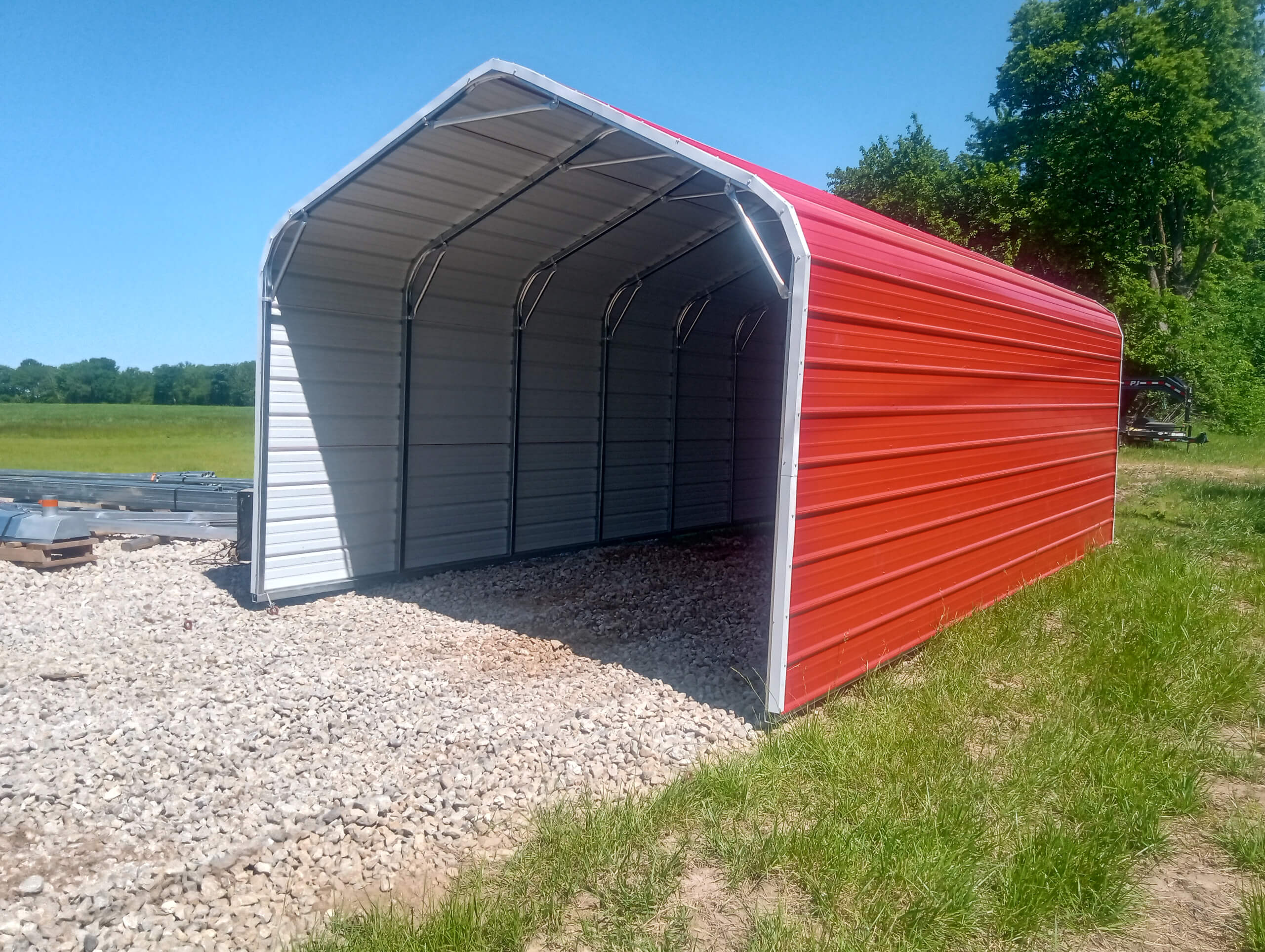 Sunny Crest Acres Versailles MO A red and gray metal carport structure stands on a gravel pad in a grassy outdoor area with trees and blue sky in the background.