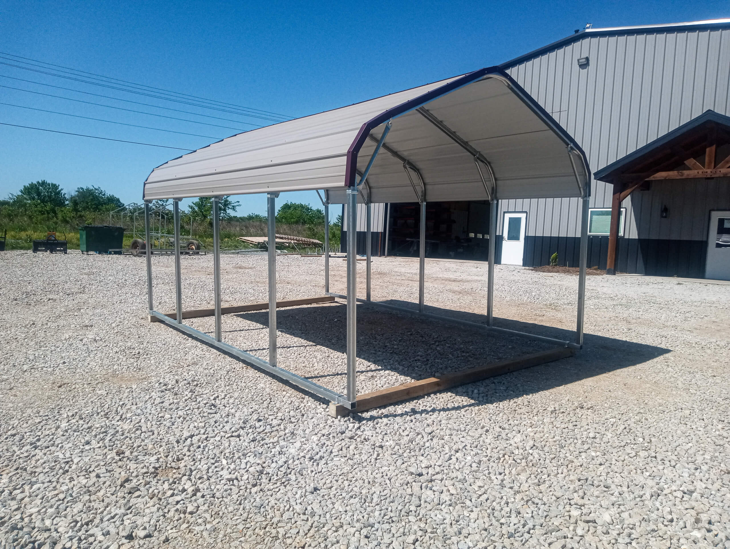 Sunny Crest Acres Versailles MO A metal carport frame with a roof but no walls or floor stands on gravel next to a gray metal building under a clear blue sky.