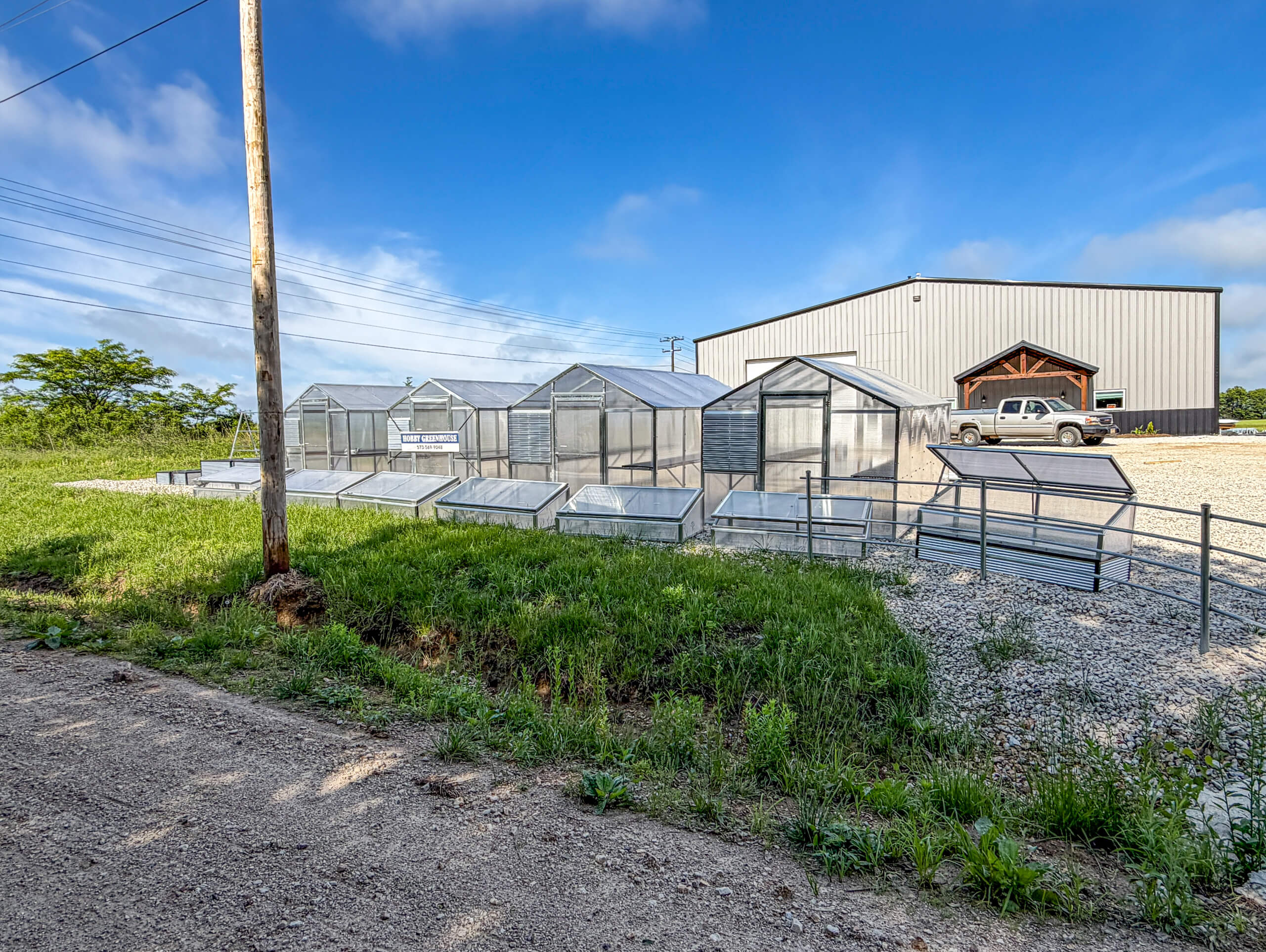 Sunny Crest Acres Versailles MO Several metal and glass greenhouses stand on a gravel lot beside a large metal building and a parked pickup truck, with grassy areas and utility lines in the background.