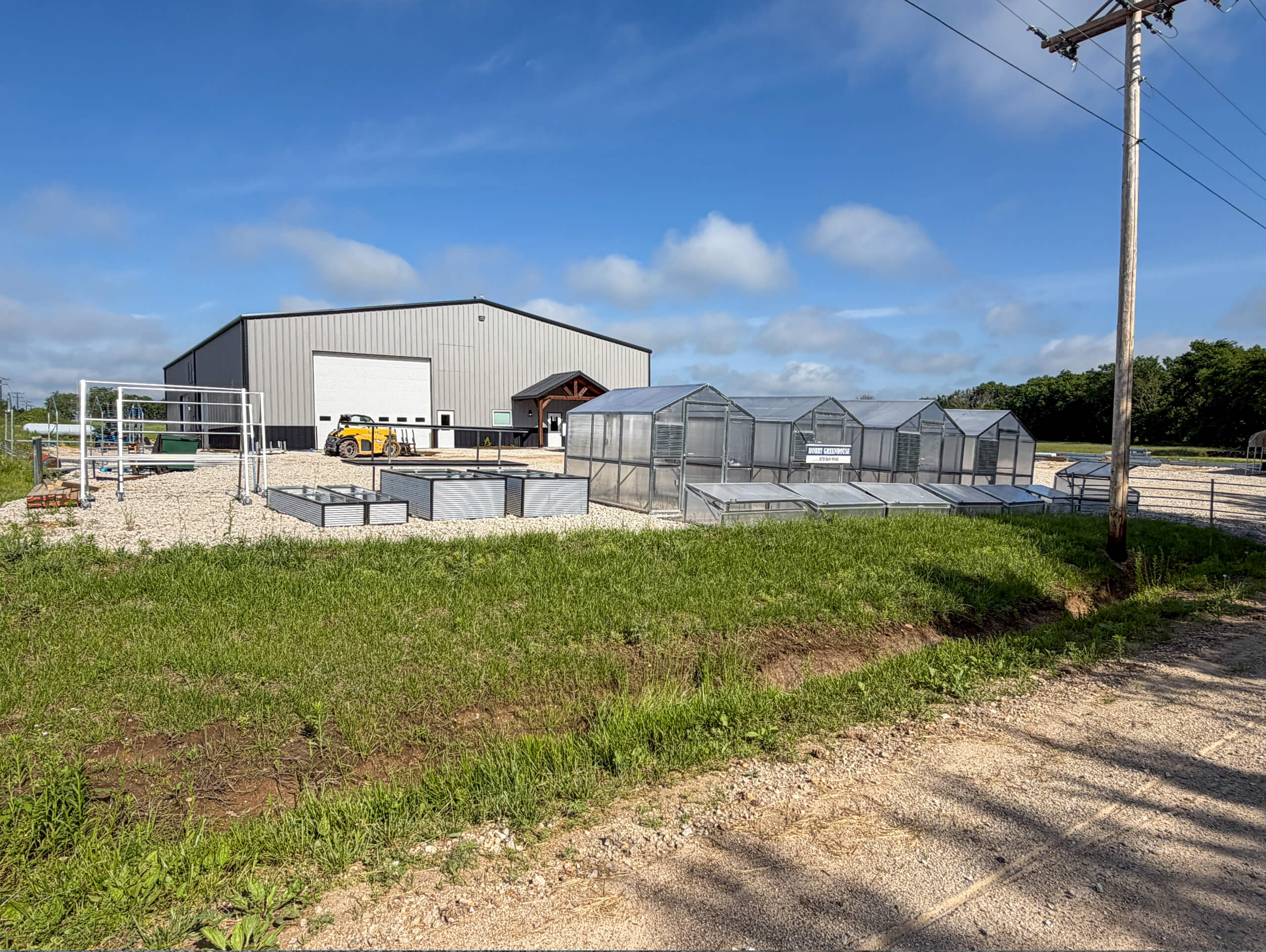 Sunny Crest Acres Versailles MO Several metal-framed greenhouses and raised garden beds sit on gravel outside a large gray industrial building on a sunny day.
