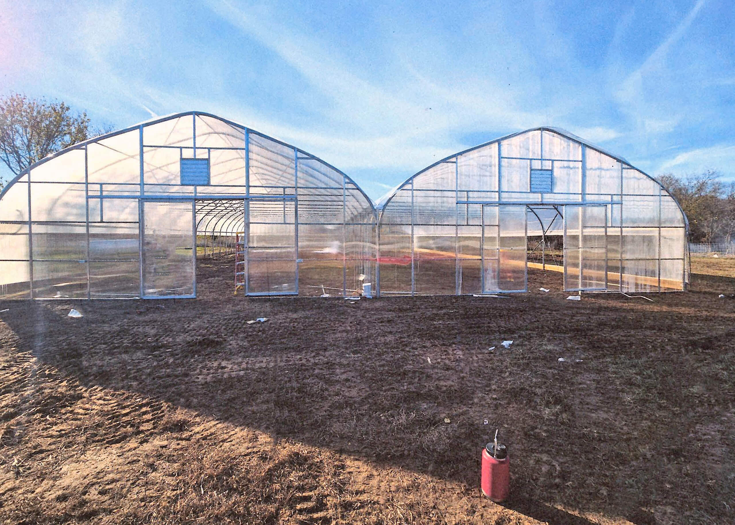 Sunny Crest Acres Versailles MO Two large, clear plastic greenhouses stand side by side on a bare dirt field under a blue sky, with a red container in the foreground.