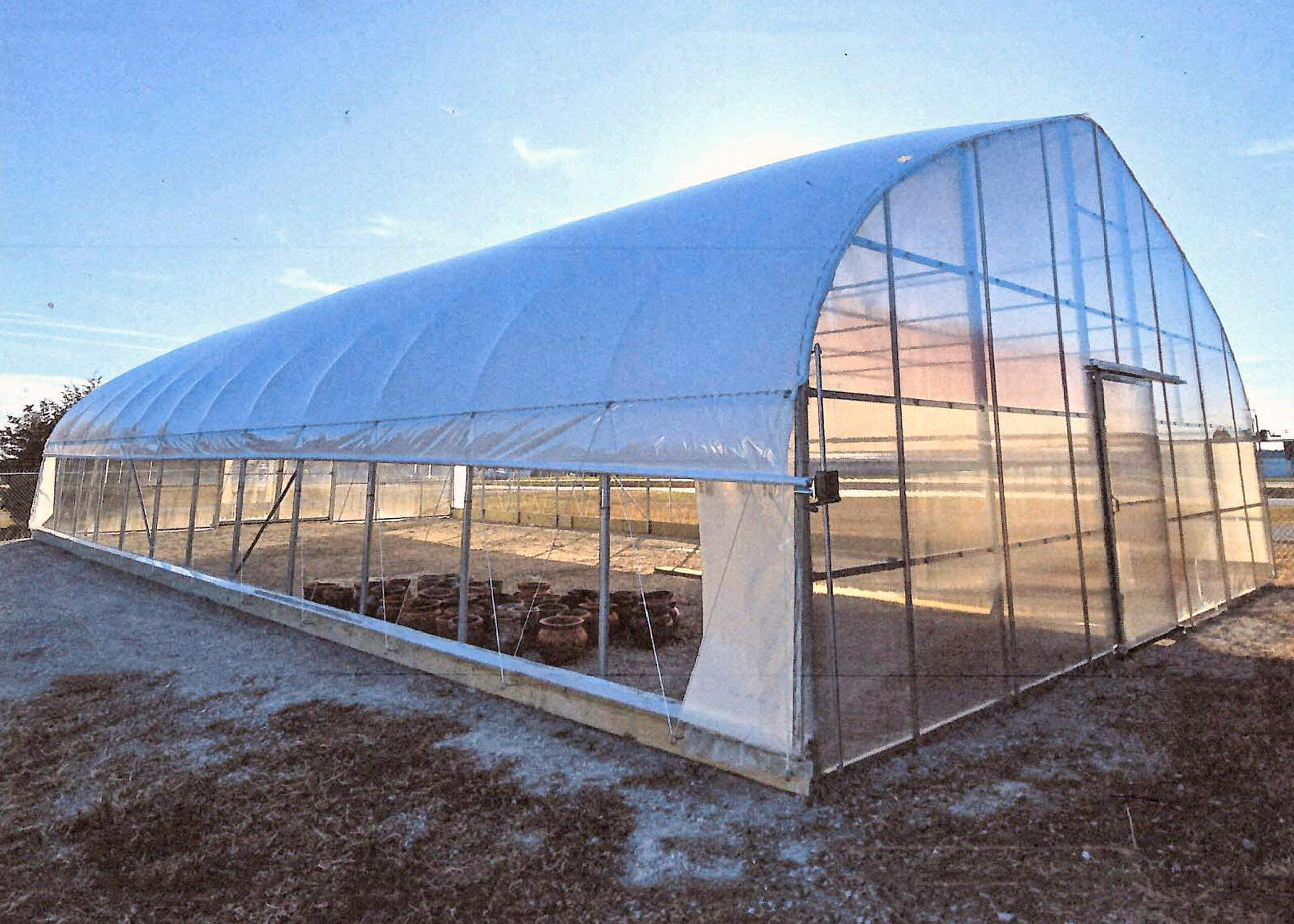 Sunny Crest Acres Versailles MO A large, empty greenhouse with a curved, translucent roof and clear plastic walls stands on a dirt lot under a blue sky.