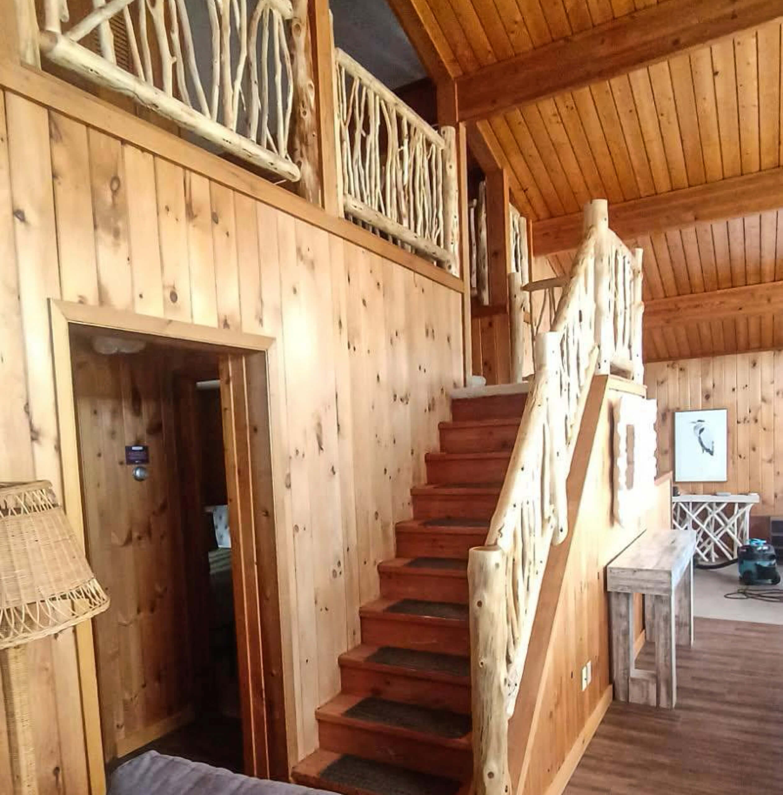Brookview Builders Penn Yan, NY Interior of a rustic wooden cabin with a staircase featuring natural branch railings, wood-paneled walls and ceiling, and simple furniture.