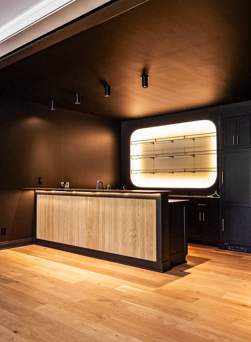Creative Cabinetry Bloomfield Iowa A modern bar area with a wooden countertop, black cabinetry, light wood flooring, and a backlit wall-mounted glass shelf.