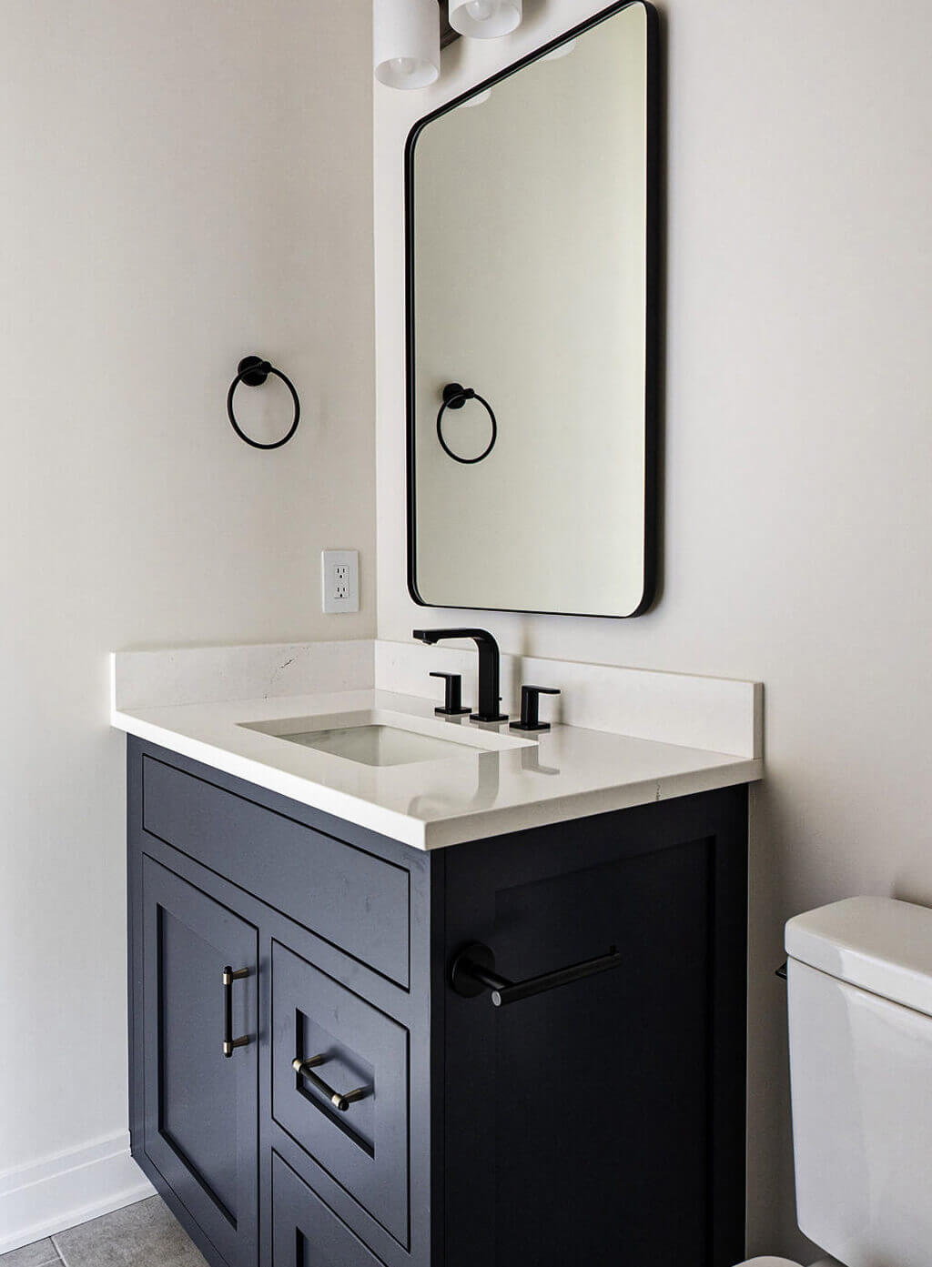 Creative Cabinetry Bloomfield Iowa A modern bathroom vanity with a black cabinet, white countertop, rectangular mirror, black faucet, and matching black hardware against a light-colored wall.