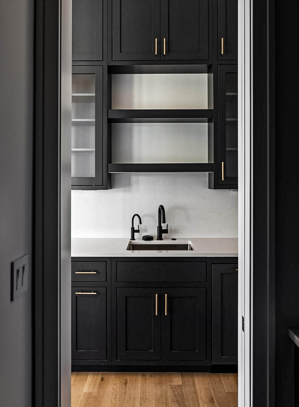 Creative Cabinetry Bloomfield Iowa Modern kitchen with matte black cabinets, gold handles, open shelves, a white countertop, black faucet, and light wood flooring.