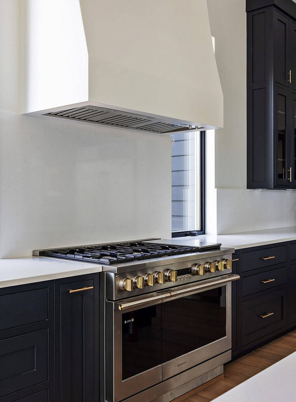 Creative Cabinetry Bloomfield Iowa Modern kitchen with a stainless steel gas range and oven, dark cabinets with brass hardware, white countertops, and a large white range hood.
