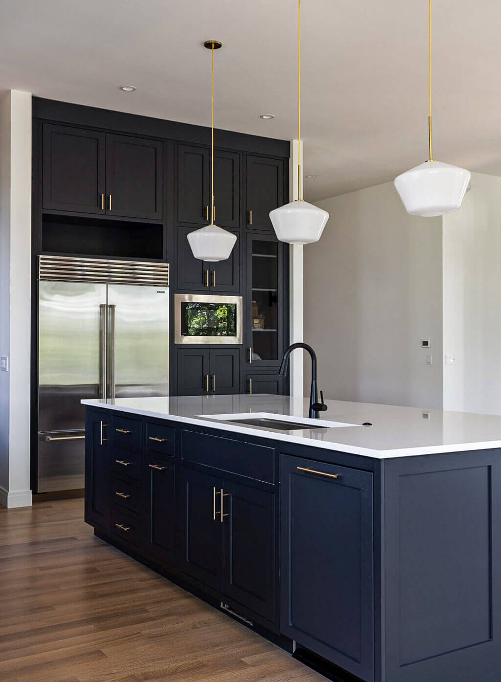 Creative Cabinetry Bloomfield Iowa Modern kitchen with dark blue cabinets, white countertops, stainless steel appliances, and three pendant lights hanging above a central island with a built-in sink.