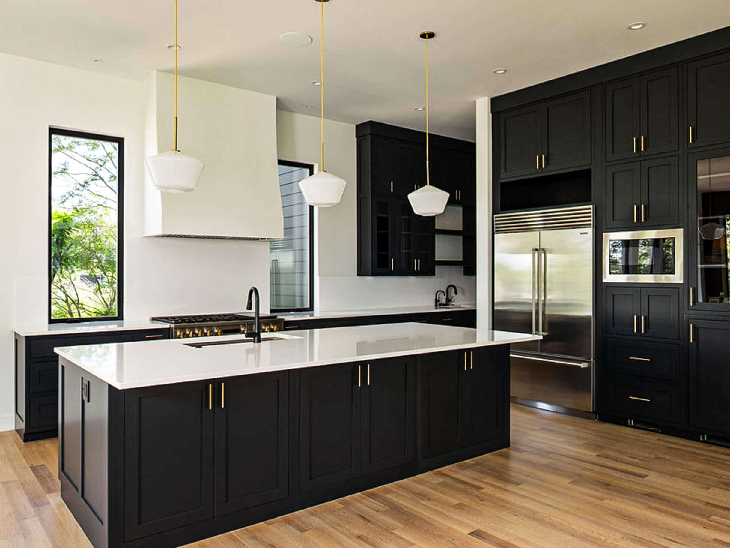 Creative Cabinetry Bloomfield Iowa Modern kitchen with black cabinets, white countertops, stainless steel appliances, three pendant lights, and hardwood floors. Large windows provide natural light.
