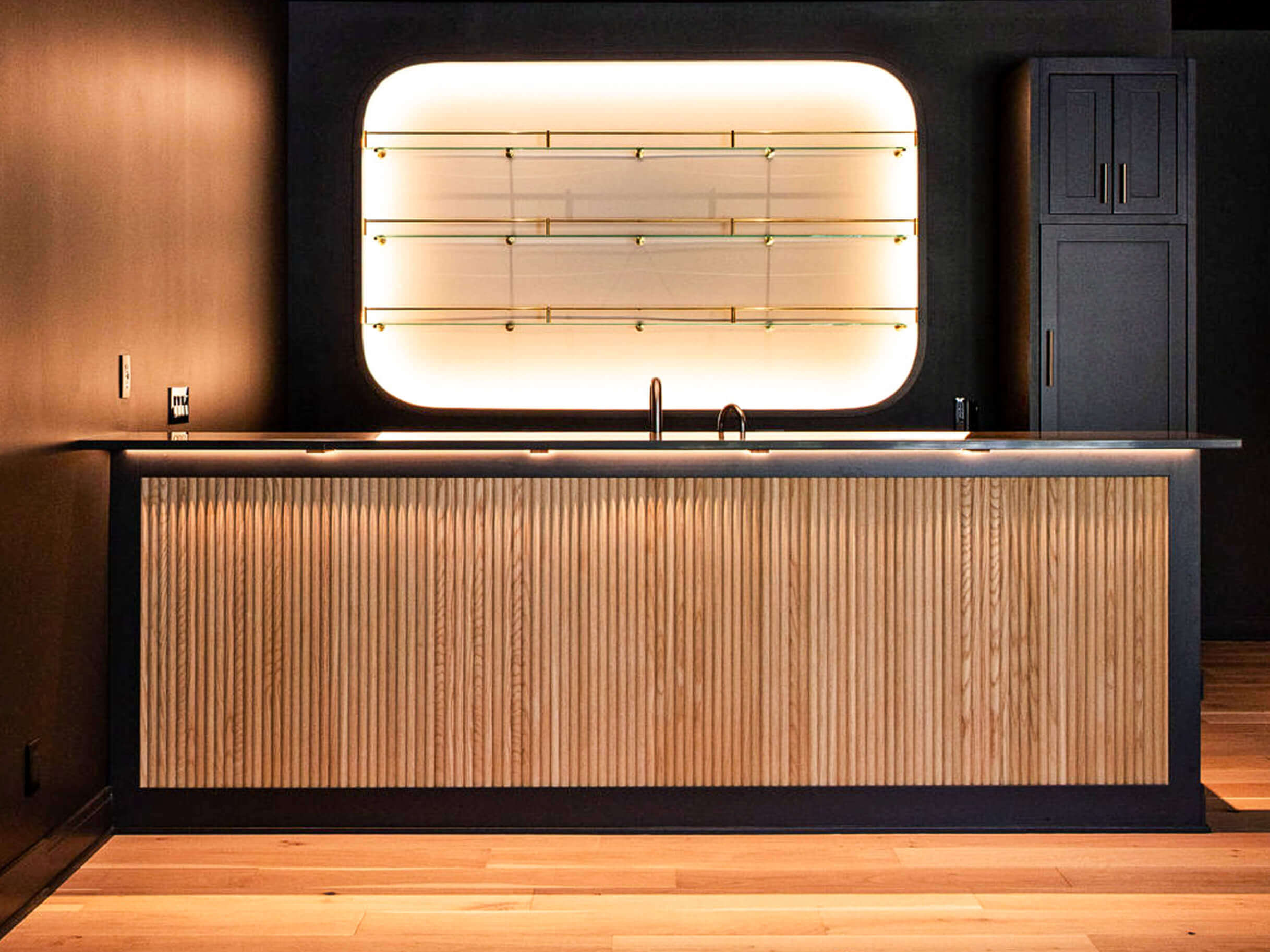 Creative Cabinetry Bloomfield Iowa A modern home bar with a ribbed wood front, black countertop, backlit glass shelves, and dark cabinetry, set against a dark wall and light wood floor.
