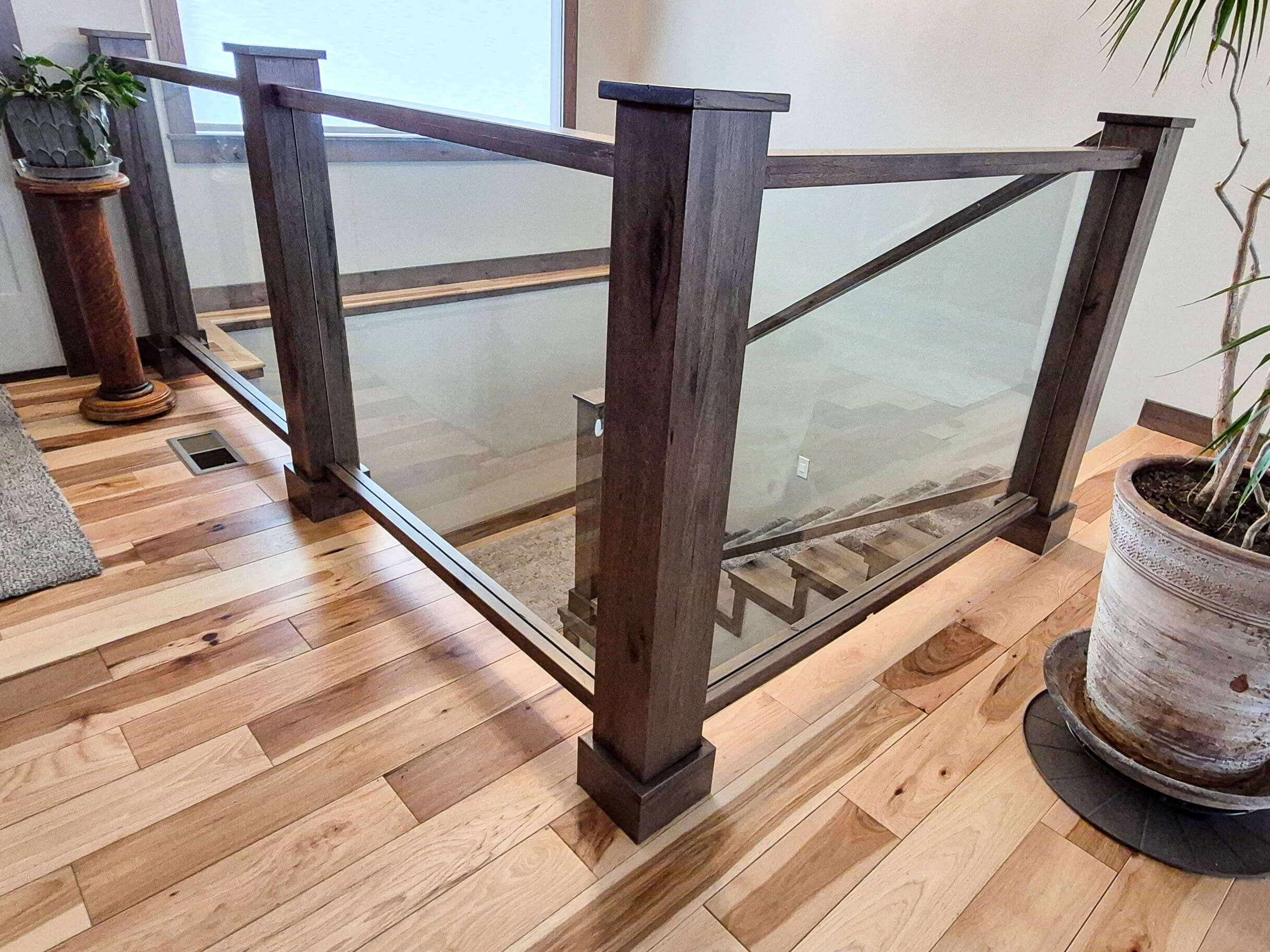 Creative Cabinetry Bloomfield Iowa A modern staircase with a glass and wood railing, set on a polished wood floor, next to potted plants and a window.