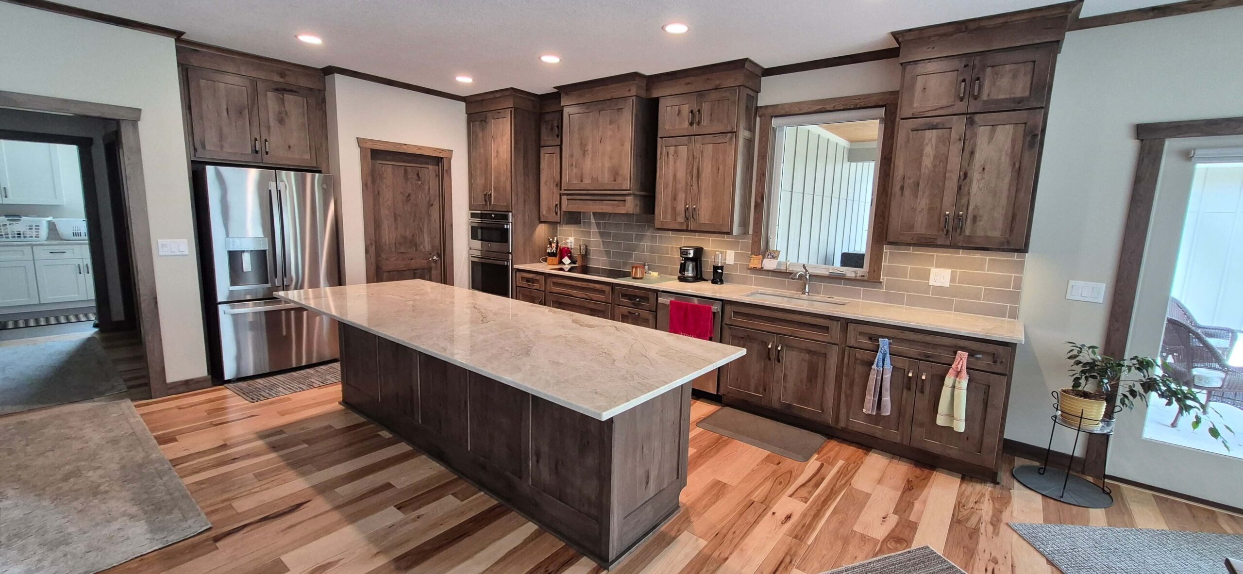 Creative Cabinetry Bloomfield Iowa Modern kitchen with wooden cabinets, stainless steel appliances, a large island with a light countertop, and hardwood flooring. Towels hang from cabinet handles; a plant sits in the corner.