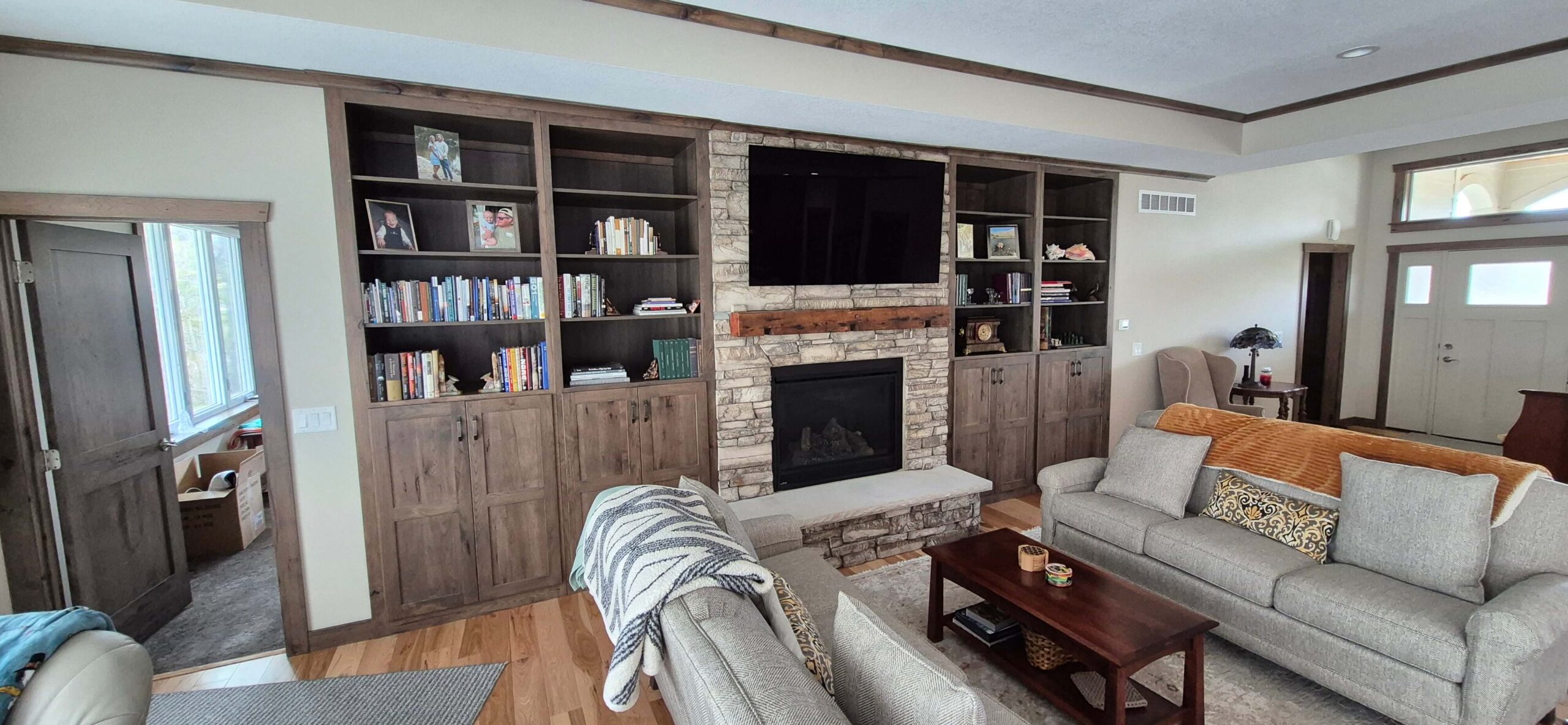 Creative Cabinetry Bloomfield Iowa A living room with a stone fireplace, a wall-mounted TV, built-in bookshelves, two beige sofas, a wooden coffee table, and a window letting in natural light.
