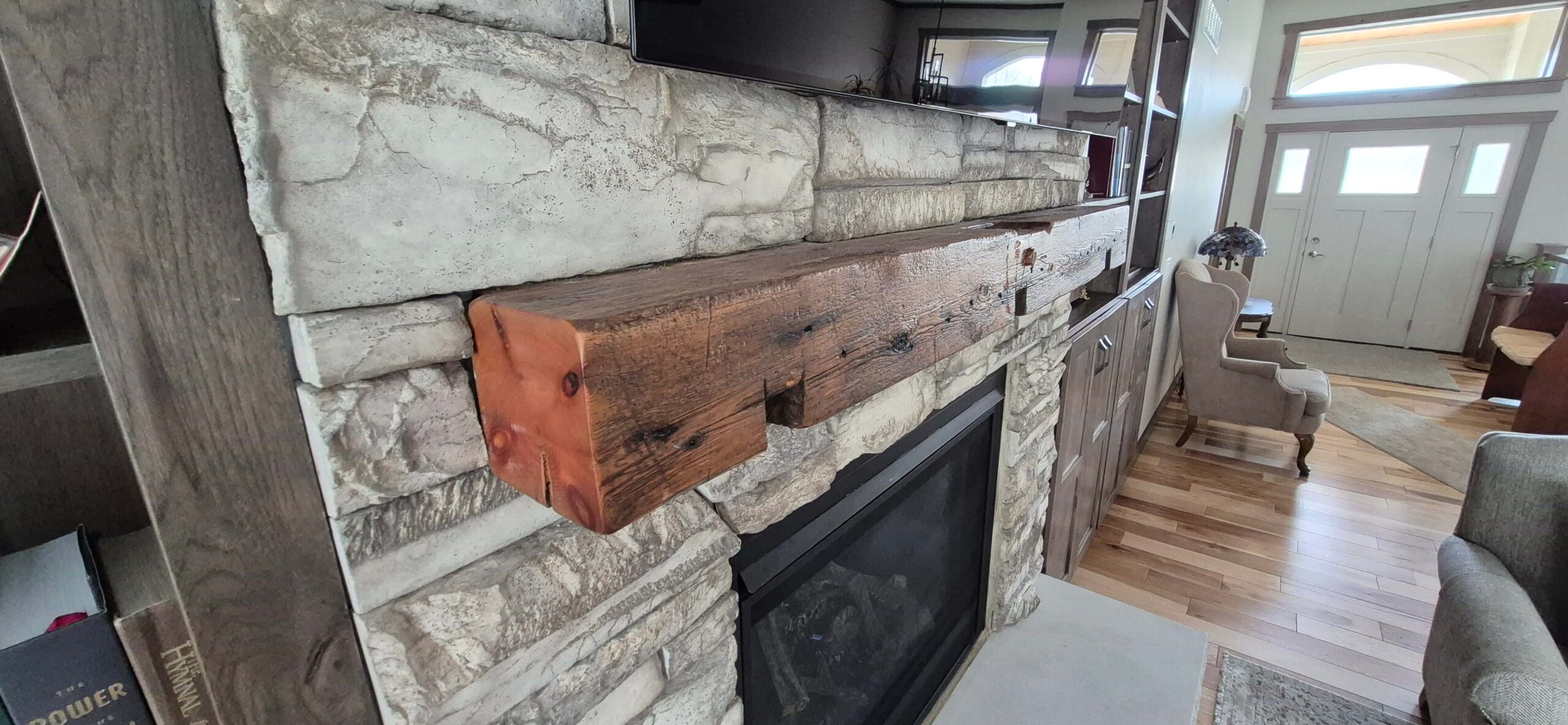 Creative Cabinetry Bloomfield Iowa A rustic wooden mantel sits above a stone fireplace in a living room with hardwood floors and built-in shelves.
