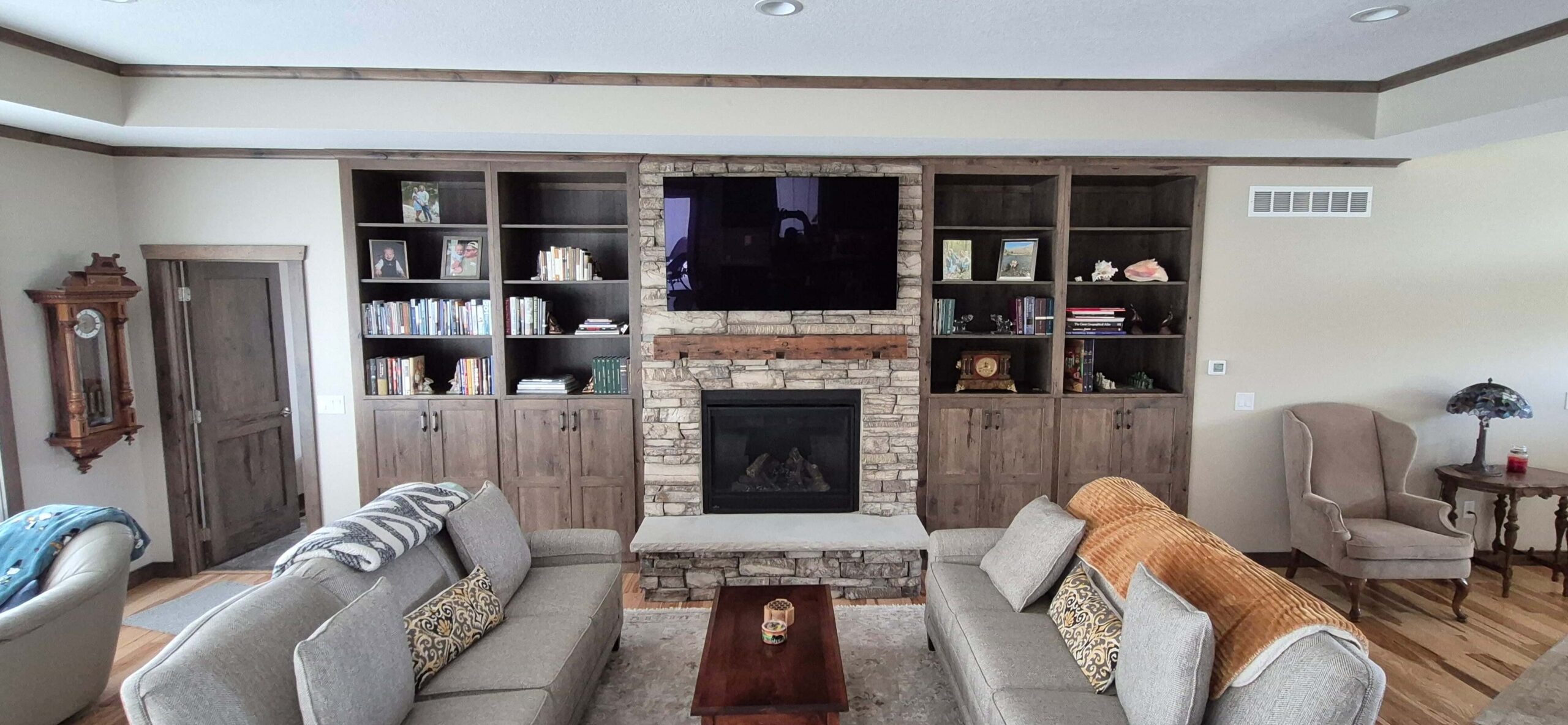 Creative Cabinetry Bloomfield Iowa Living room with two sofas, a stone fireplace, a wall-mounted TV, built-in shelves with books and decor, and a grandfather clock on the left.