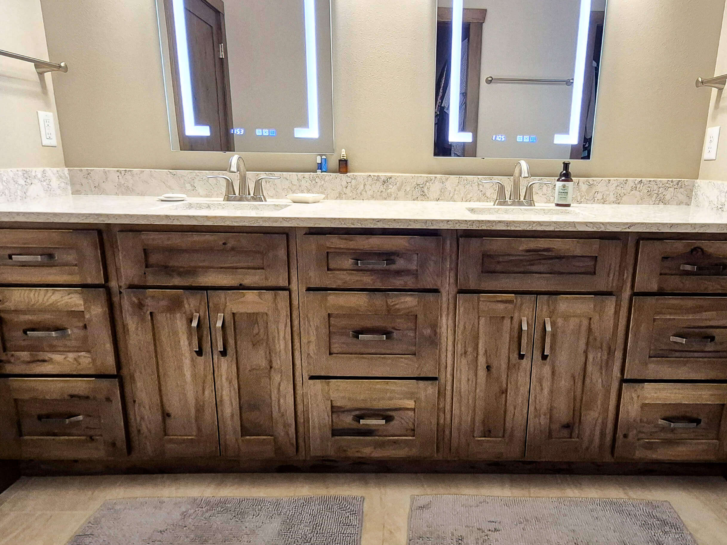 Creative Cabinetry Bloomfield Iowa Double-sink bathroom vanity with wooden cabinets, marble countertop, two mirrors with LED lighting, and minimal toiletries on the counter.