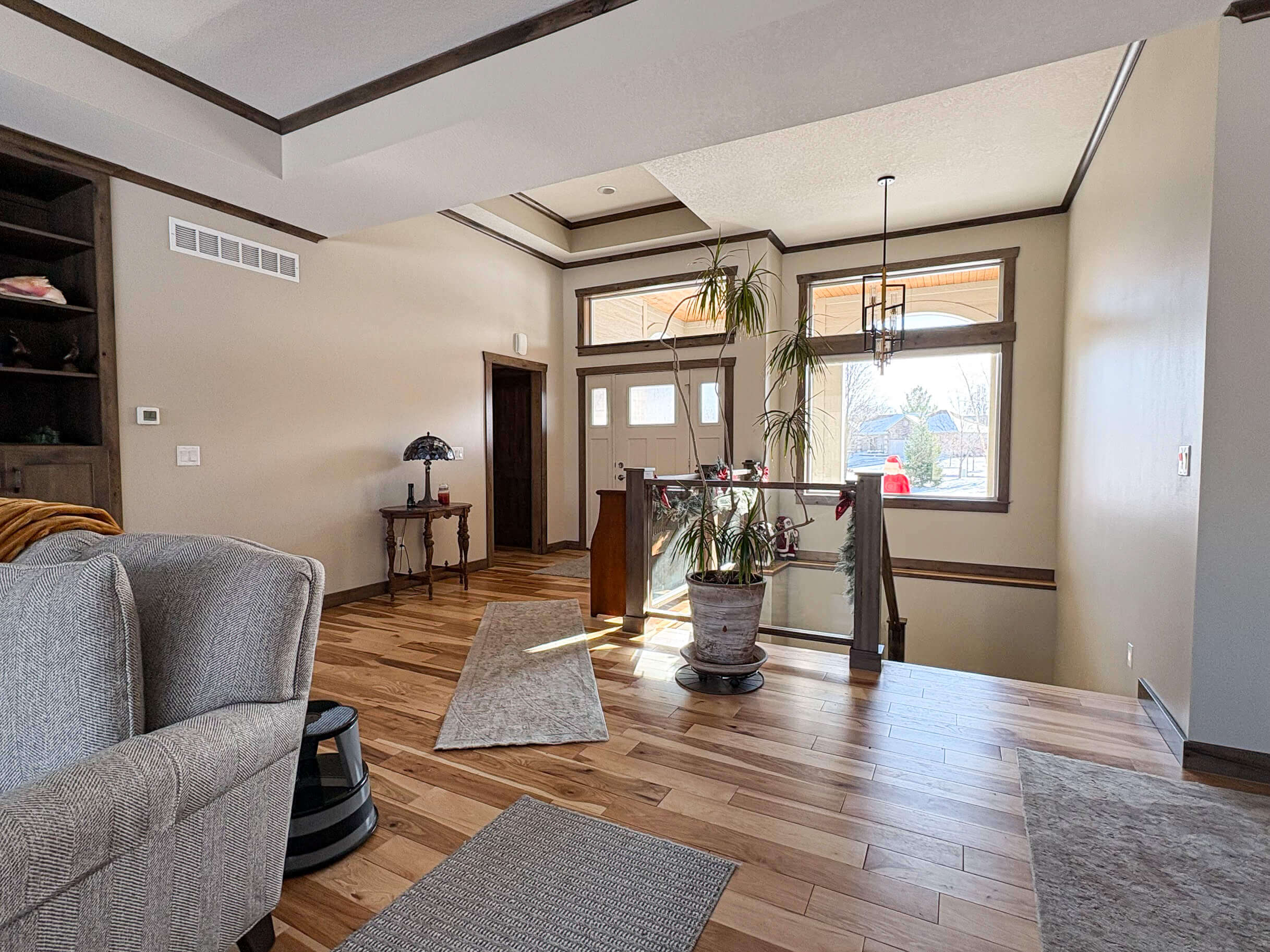 Creative Cabinetry Bloomfield Iowa A spacious, well-lit entryway with wooden floors, a potted plant, a small table with a lamp, and stairs leading down; sunlight streams through large front windows.