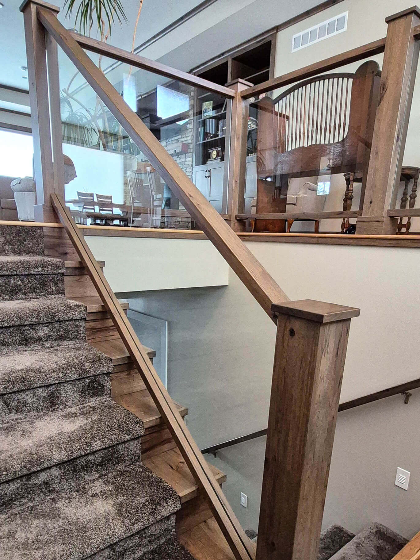 Creative Cabinetry Bloomfield Iowa Wooden staircase with glass panel railing and carpeted steps, leading to an upper level with wooden furniture visible in the background.