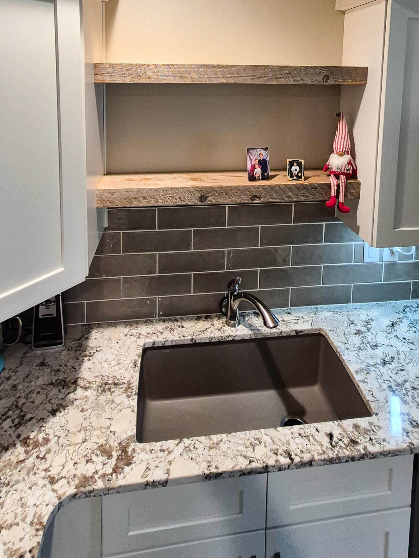 Creative Cabinetry Bloomfield Iowa A kitchen sink with a marbled countertop, gray tile backsplash, wooden shelves, and small decorative items, including a gnome figure, on the shelf above the sink.