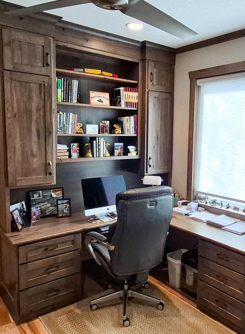 Creative Cabinetry Bloomfield Iowa A home office with a wooden desk, a black office chair, built-in shelves holding books and decor, and a large computer monitor near a window with natural light.