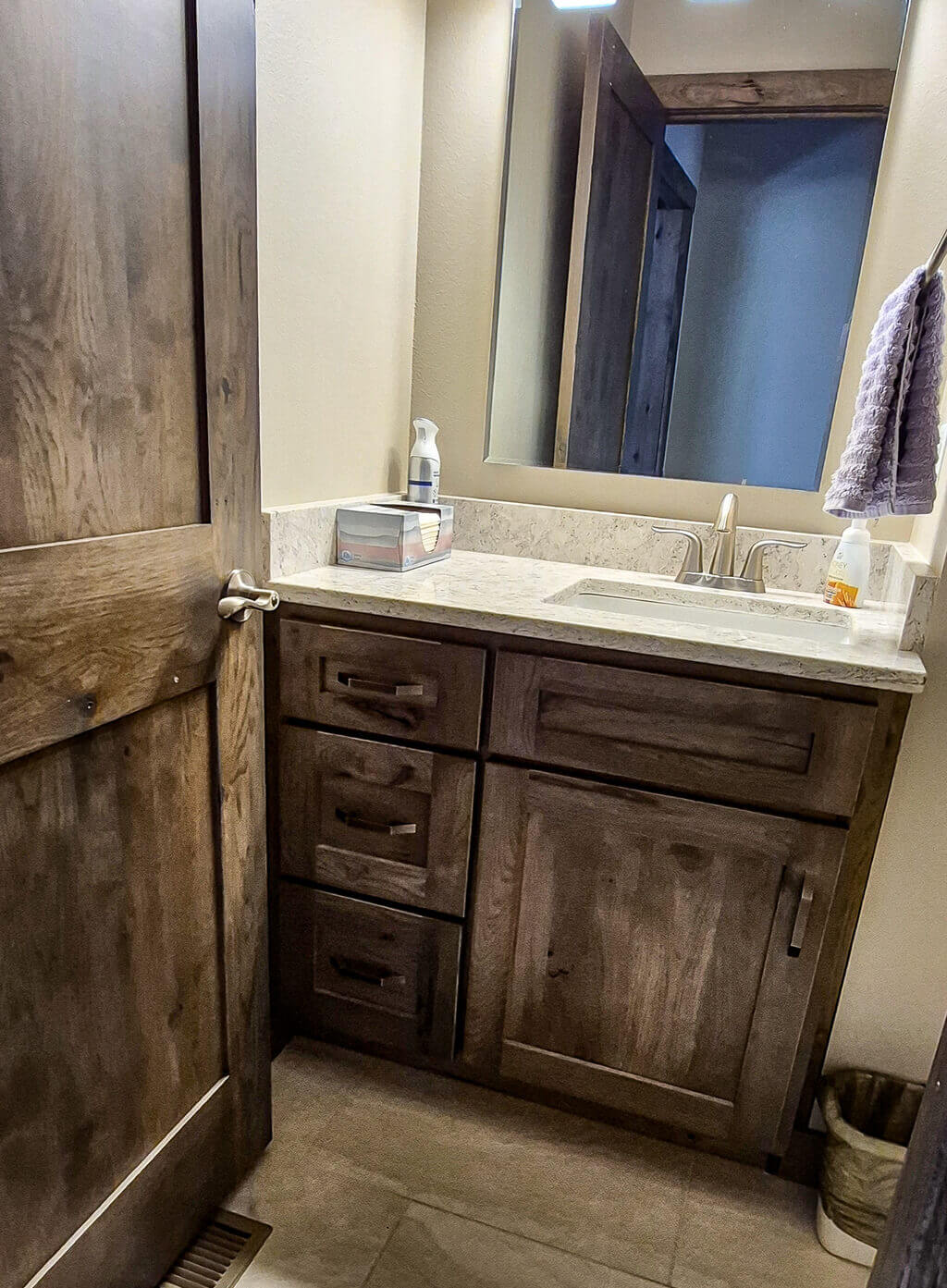 Creative Cabinetry Bloomfield Iowa A bathroom with a wooden vanity, marble countertop, sink, mirror, soap dispenser, tissue box, towel on a rack, and a trash bin on the floor.
