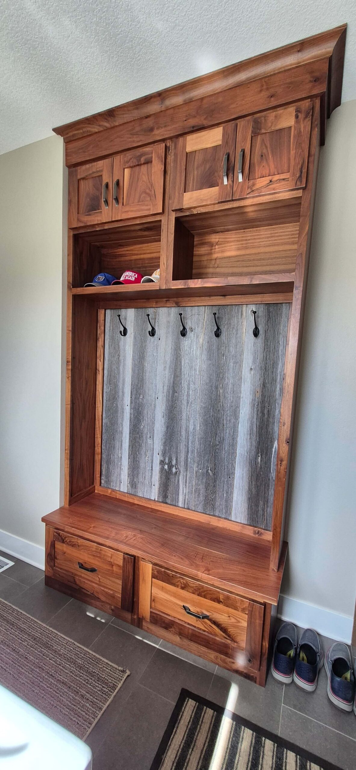 Creative Cabinetry Bloomfield Iowa A wooden hall tree with upper cabinets, cubbies holding hats, five coat hooks, a wooden bench, two lower drawers, and shoes placed to the right on the floor.