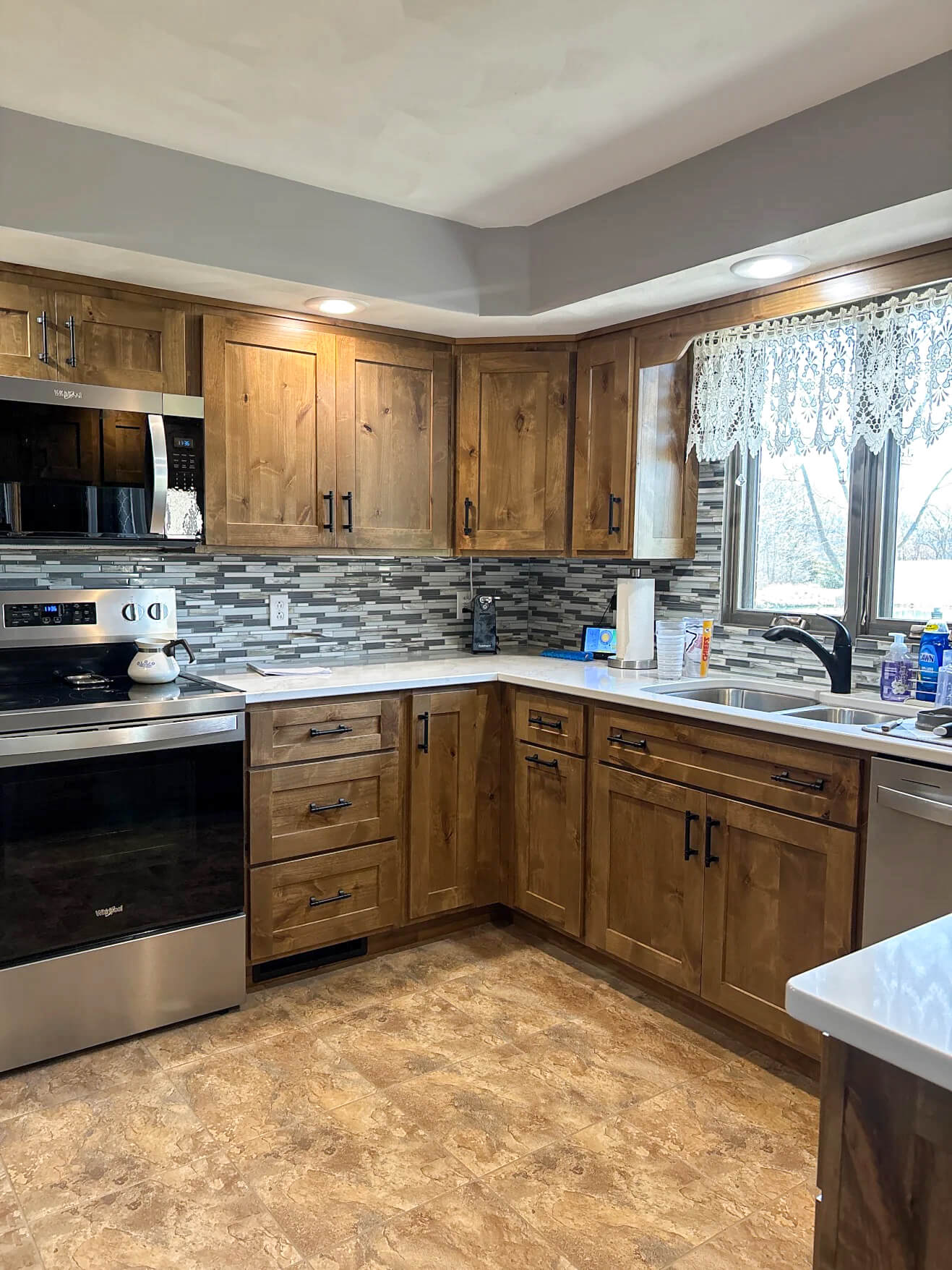 Creative Cabinetry Bloomfield Iowa Modern kitchen with wooden cabinets, stainless steel appliances, tiled backsplash, and a window with lace curtains above the sink.