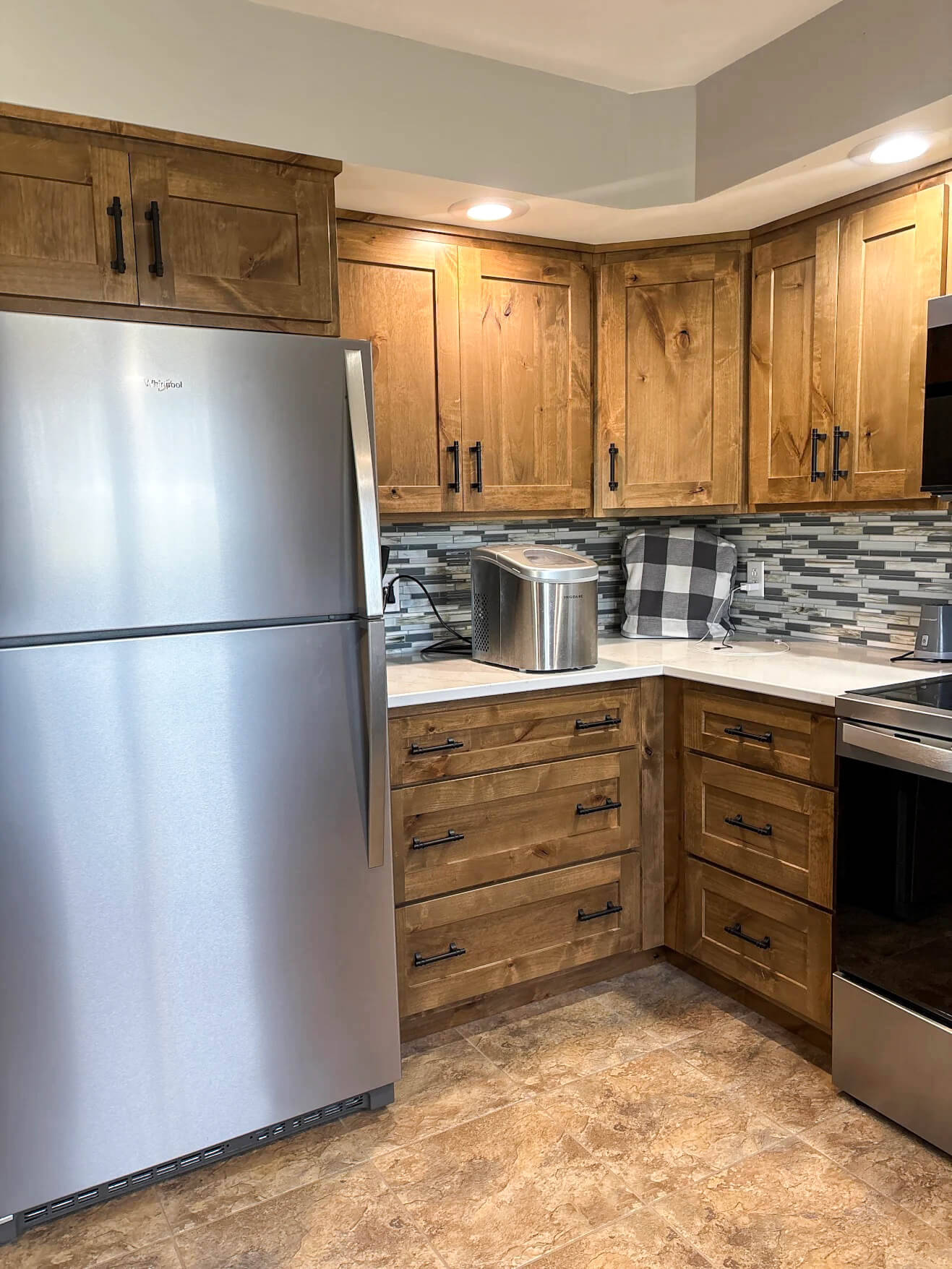 Creative Cabinetry Bloomfield Iowa Modern kitchen with stainless steel refrigerator, wooden cabinets, a metal bread box, and a checkered kitchen towel on the countertop.