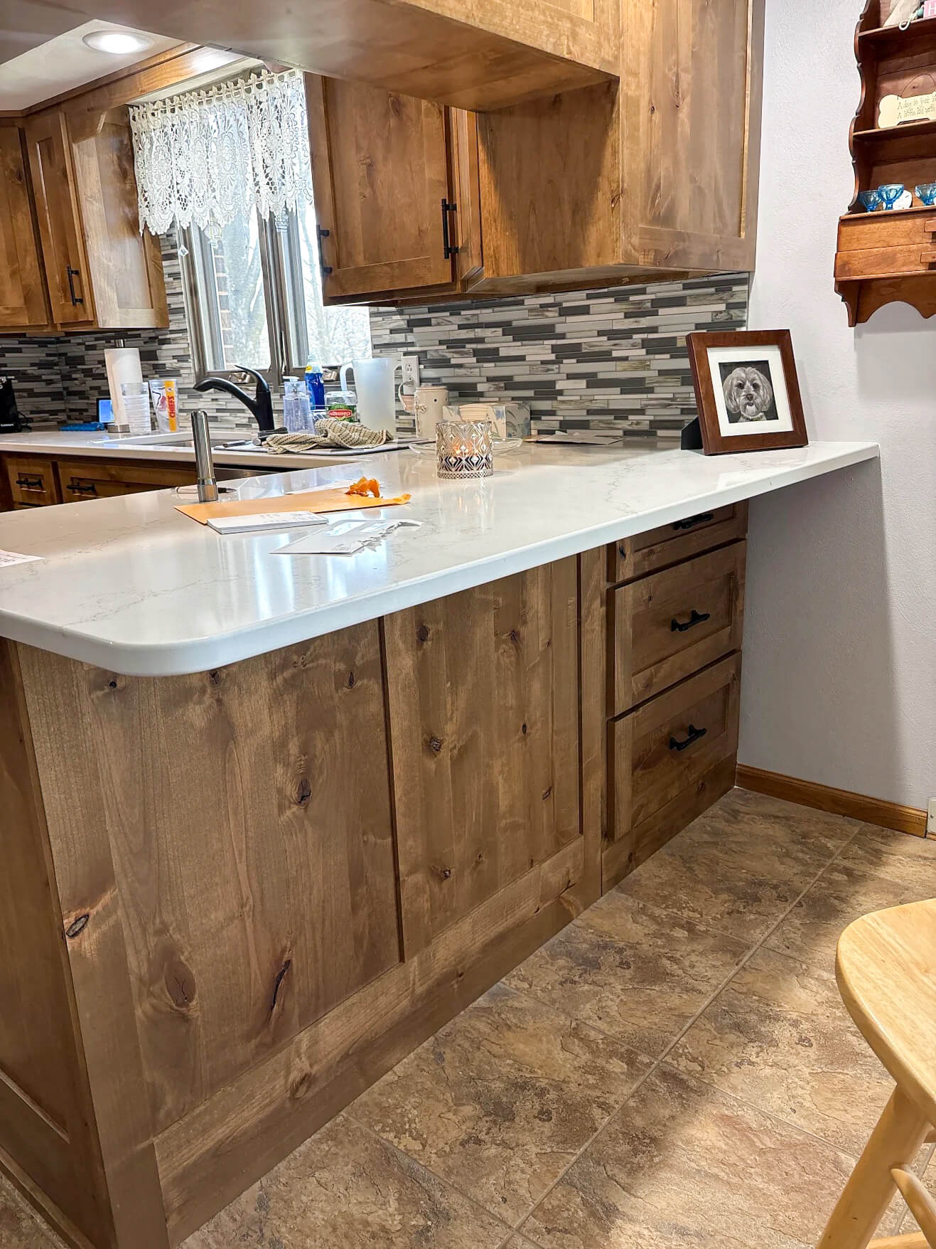 Creative Cabinetry Bloomfield Iowa A wooden kitchen with a white countertop, mosaic tile backsplash, sink, various items on the counter, and a framed photo on the ledge.