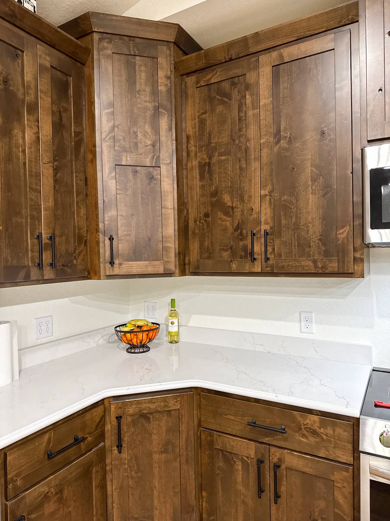 Creative Cabinetry Bloomfield Iowa Kitchen corner with dark wood cabinets, white marble countertop, a fruit bowl, a bottle of wine, and a paper towel roll.