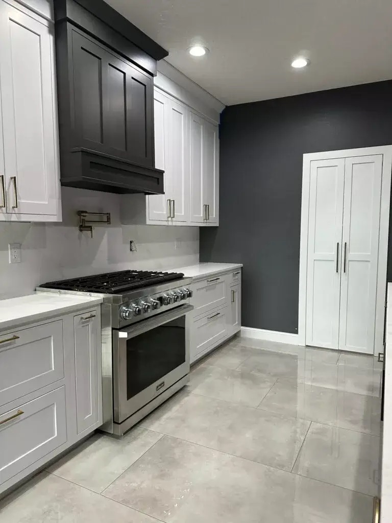 Creative Cabinetry Bloomfield Iowa Modern kitchen with white cabinets, a stainless steel gas range, dark vent hood, light tile flooring, and a gray accent wall with double pantry doors.