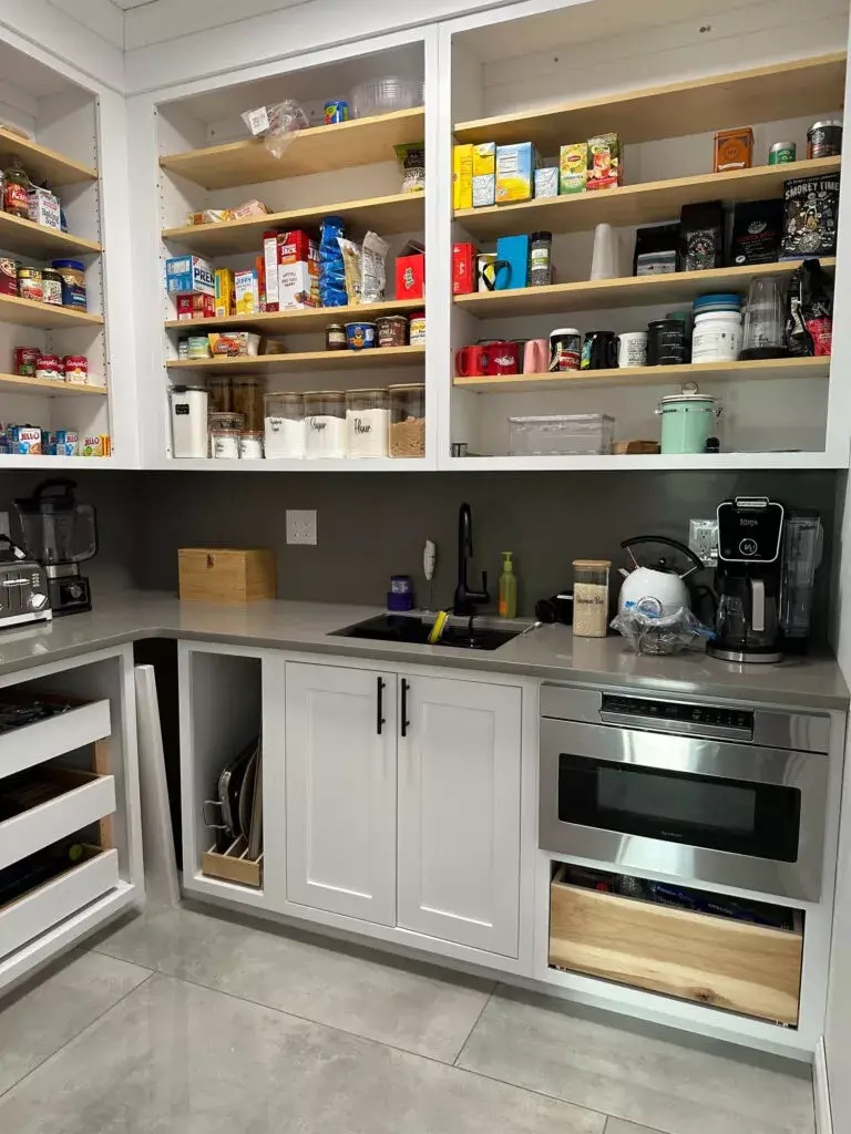Creative Cabinetry Bloomfield Iowa A modern kitchen pantry with open shelves stocked with food items and appliances, a sink, coffee machine, and closed drawers and cabinets below the countertop.