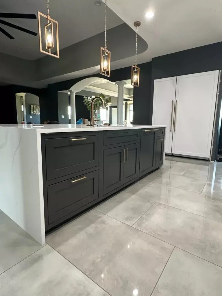 Creative Cabinetry Bloomfield Iowa Modern kitchen with gray cabinets, brass handles, white marble countertops, pendant lights, and a large refrigerator on polished tile flooring.