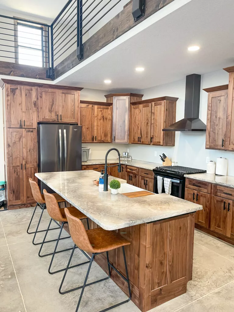 Creative Cabinetry Bloomfield Iowa Modern kitchen with wood cabinets, a marble island with three brown chairs, stainless steel appliances, and open railing on an upper level.