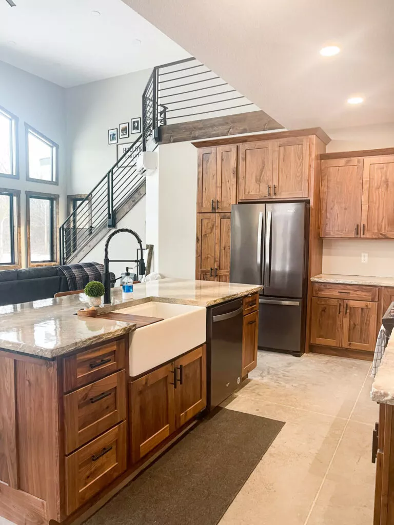Creative Cabinetry Bloomfield Iowa Modern kitchen with wooden cabinets, a farmhouse sink, stainless steel appliances, and a central island; open stairway and large windows in the background.