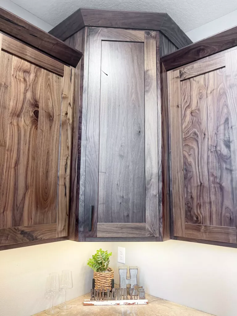 Creative Cabinetry Bloomfield Iowa Three wooden kitchen cabinets in a corner, with natural wood grain visible. Countertop below holds a small potted plant and glassware.
