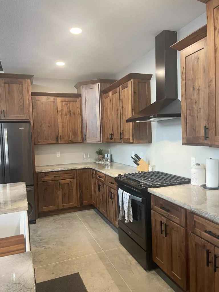 Creative Cabinetry Bloomfield Iowa Modern kitchen with wooden cabinets, stainless steel refrigerator, black stove with range hood, marble countertops, and tile flooring. Cooking utensils and decor are visible on the countertop.
