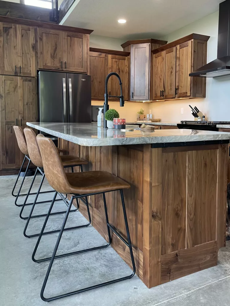 Creative Cabinetry Bloomfield Iowa Modern kitchen with wooden cabinets, a large island with a marble countertop, four brown barstools, a black faucet, and stainless steel appliances.