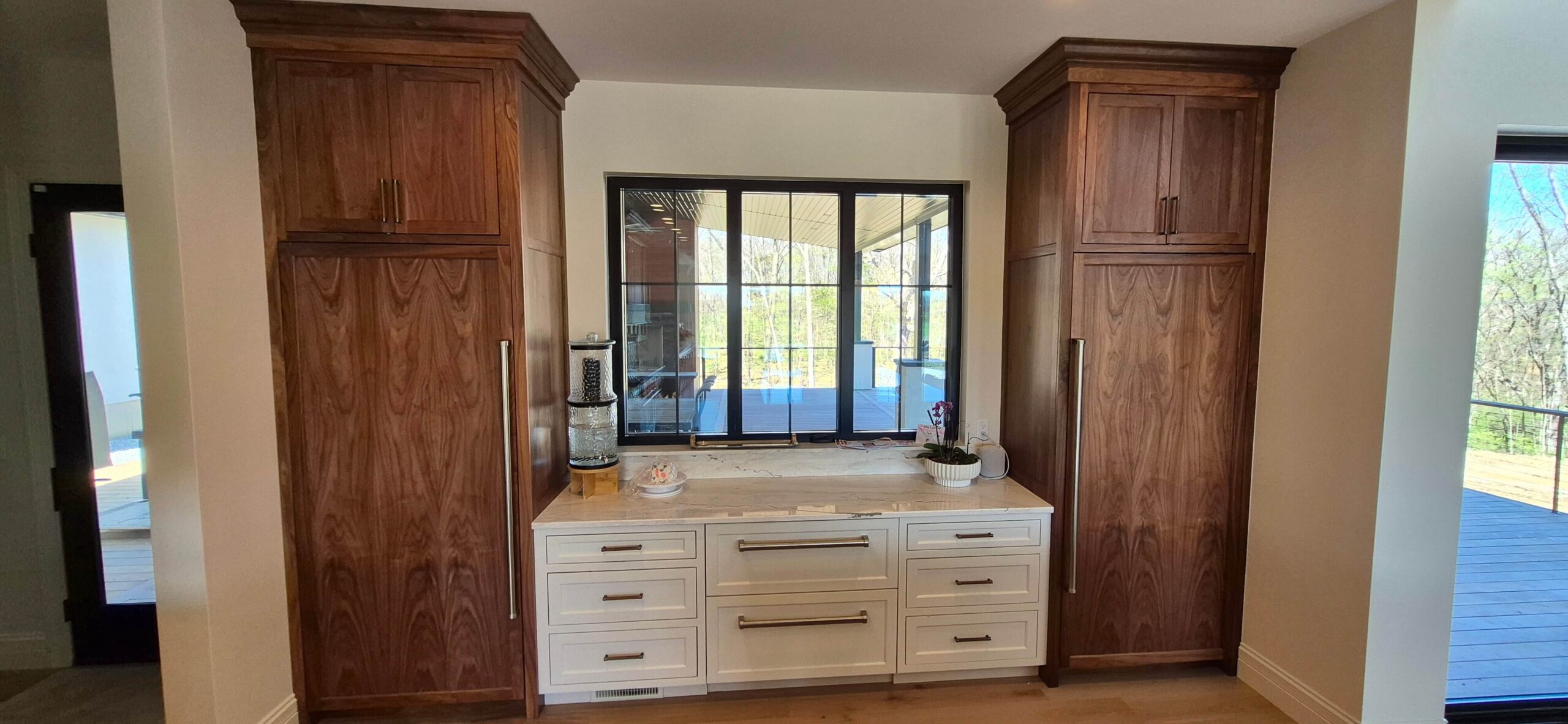 Creative Cabinetry Bloomfield Iowa A modern kitchen area with two tall wooden cabinets, a white countertop, built-in drawers, and a large window overlooking a deck and trees outside.