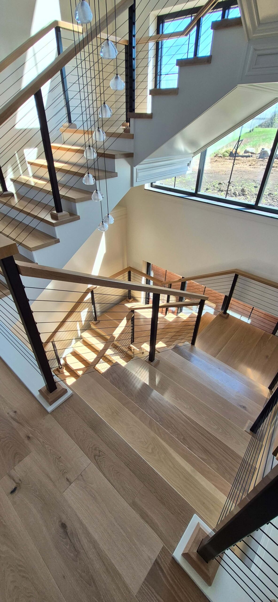 Creative Cabinetry Bloomfield Iowa Wooden staircase with metal railings and cable wires, illuminated by natural light from large windows; multiple landings and modern pendant lights visible.