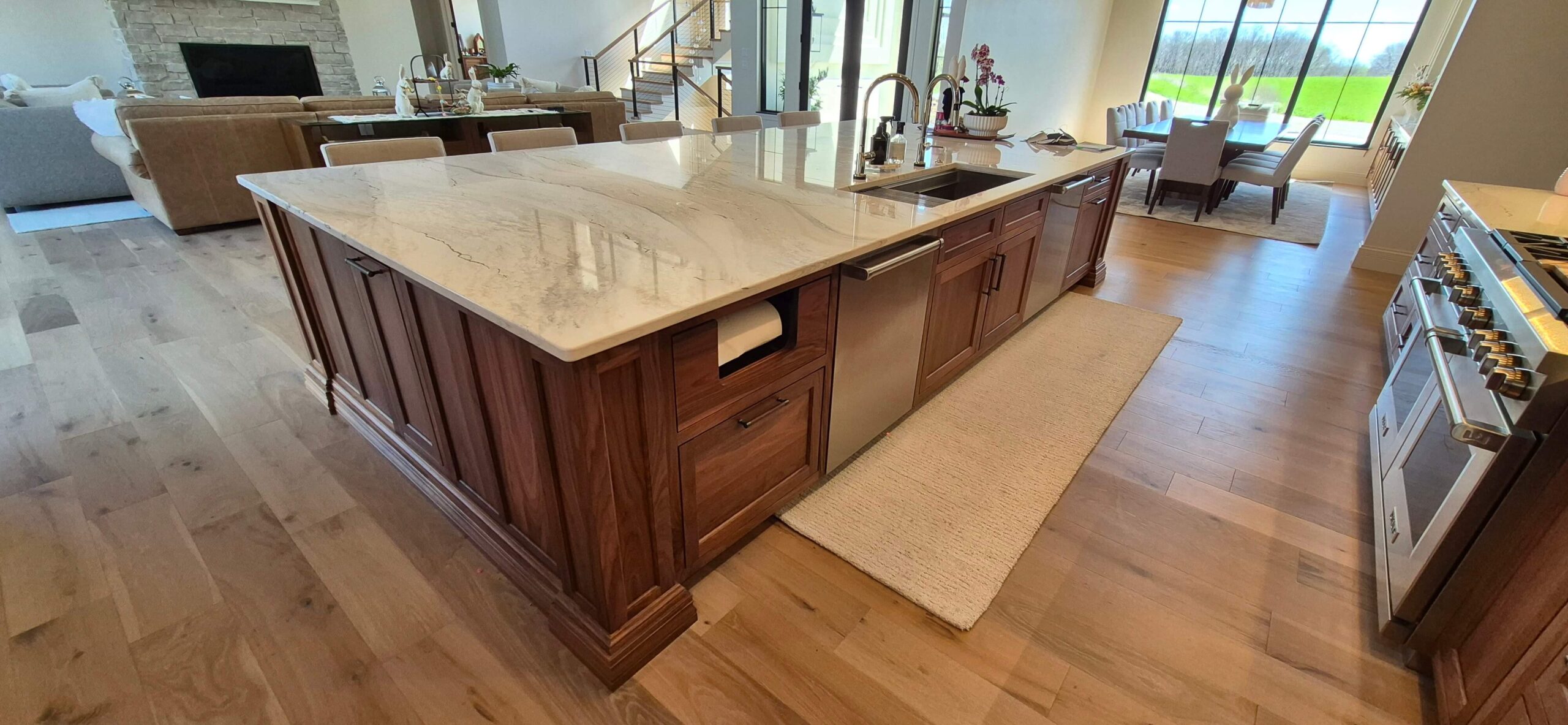 Creative Cabinetry Bloomfield Iowa A large kitchen island with a marble countertop, sink, and wood cabinetry sits in an open-plan living space with hardwood floors and natural light from large windows.