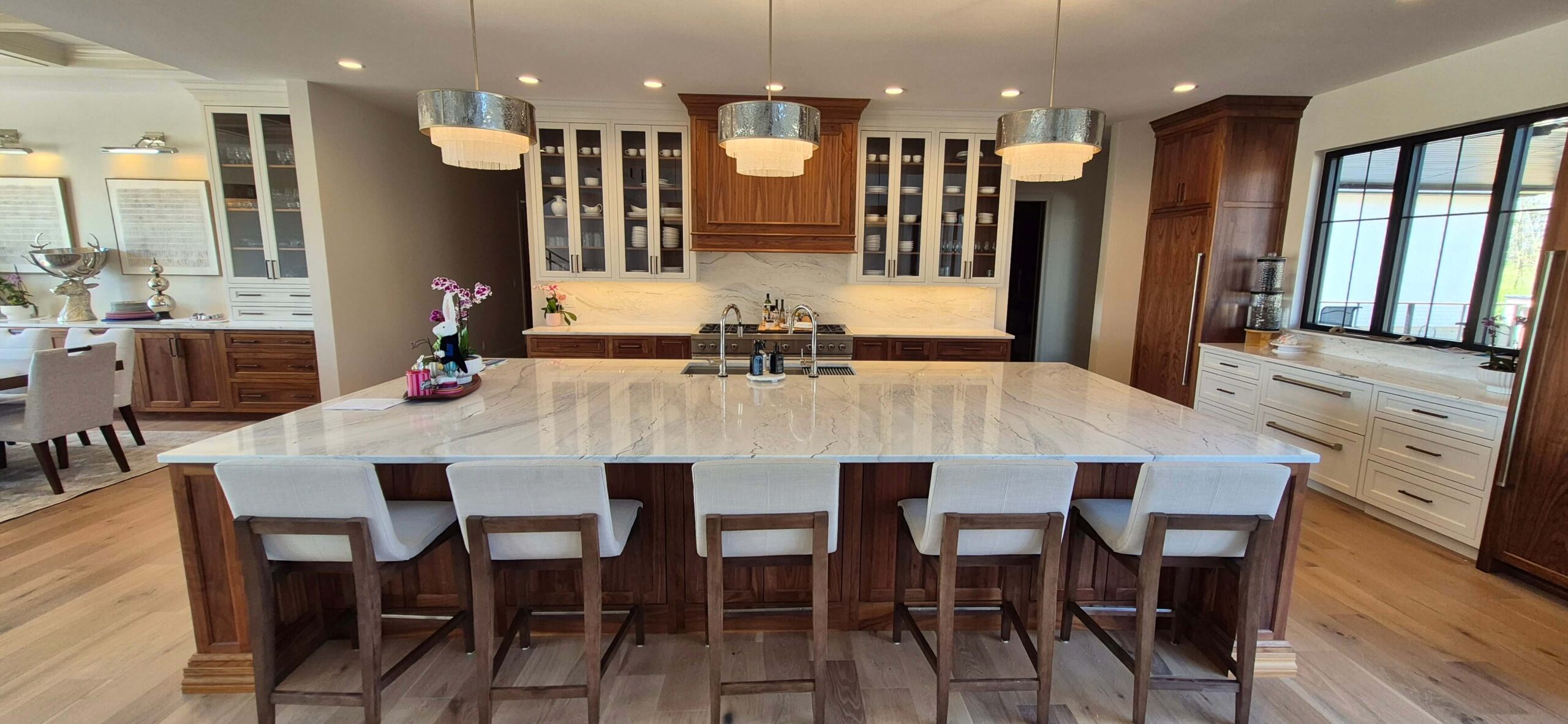 Creative Cabinetry Bloomfield Iowa Modern kitchen with a large marble island, five upholstered bar stools, wood cabinetry, glass-front cabinets, and three pendant lights above the island.