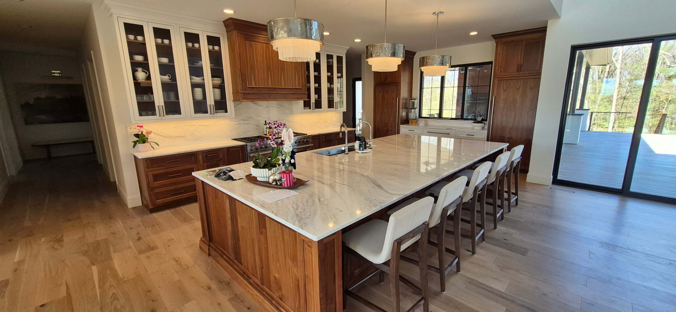 Creative Cabinetry Bloomfield Iowa Spacious kitchen with a large marble island, wooden cabinets, three pendant lights, bar stools, and floor-to-ceiling windows overlooking a deck.
