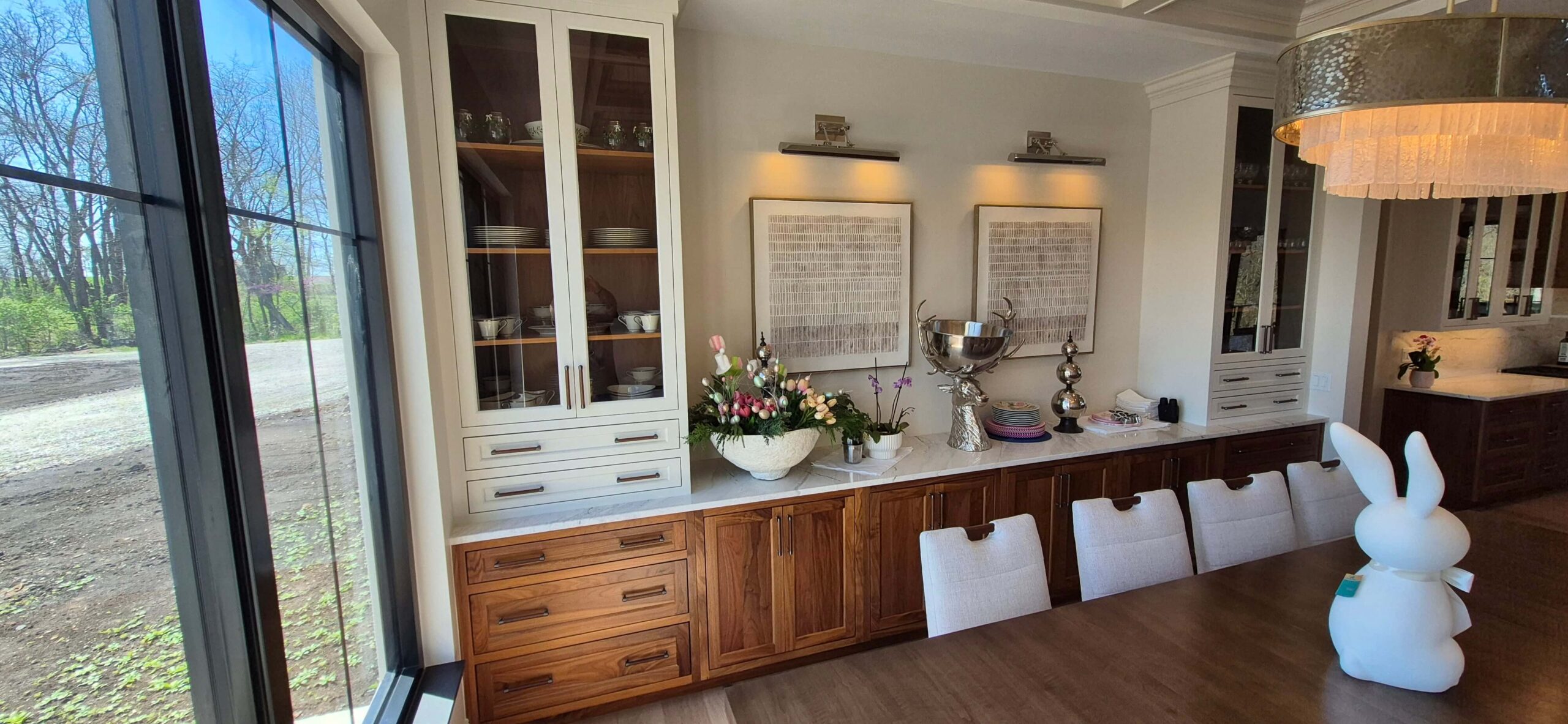 Creative Cabinetry Bloomfield Iowa Modern dining room with a wooden table, white chairs, a sideboard displaying decorative items, and large windows showing an outdoor view.