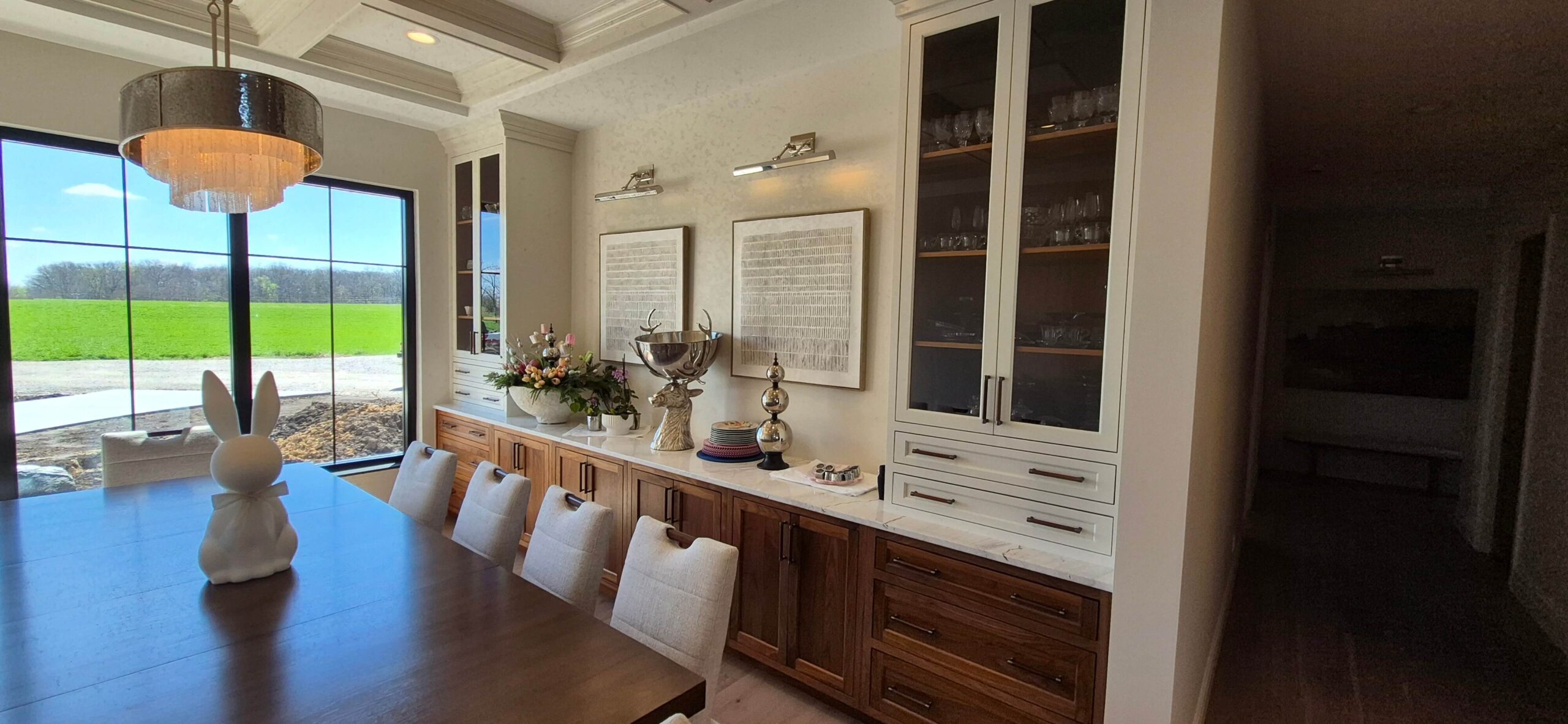 Creative Cabinetry Bloomfield Iowa A dining room with a wooden table, white chairs, a bunny-shaped centerpiece, built-in cabinets, wall art, and a large window overlooking a green field.