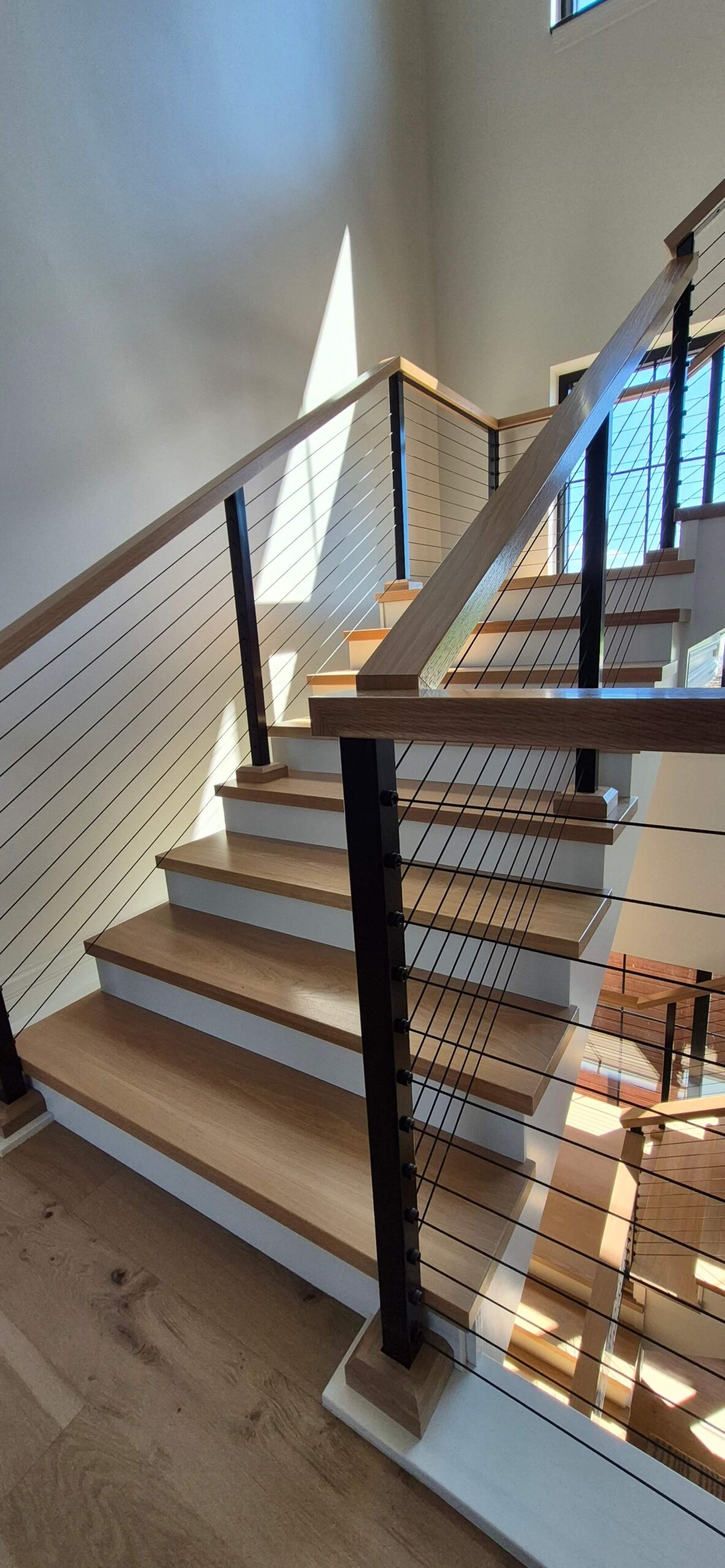 Creative Cabinetry Bloomfield Iowa A modern wooden staircase with black metal railings and cable wires, illuminated by natural sunlight through nearby windows.
