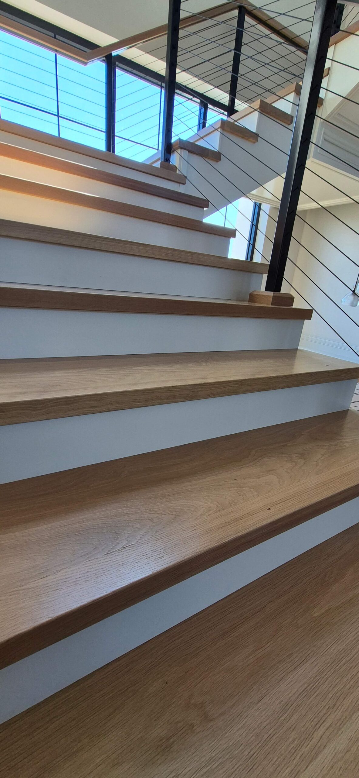 Creative Cabinetry Bloomfield Iowa Close-up view of modern wooden stairs with white risers and cable railing, leading to an upper floor with large windows in the background.