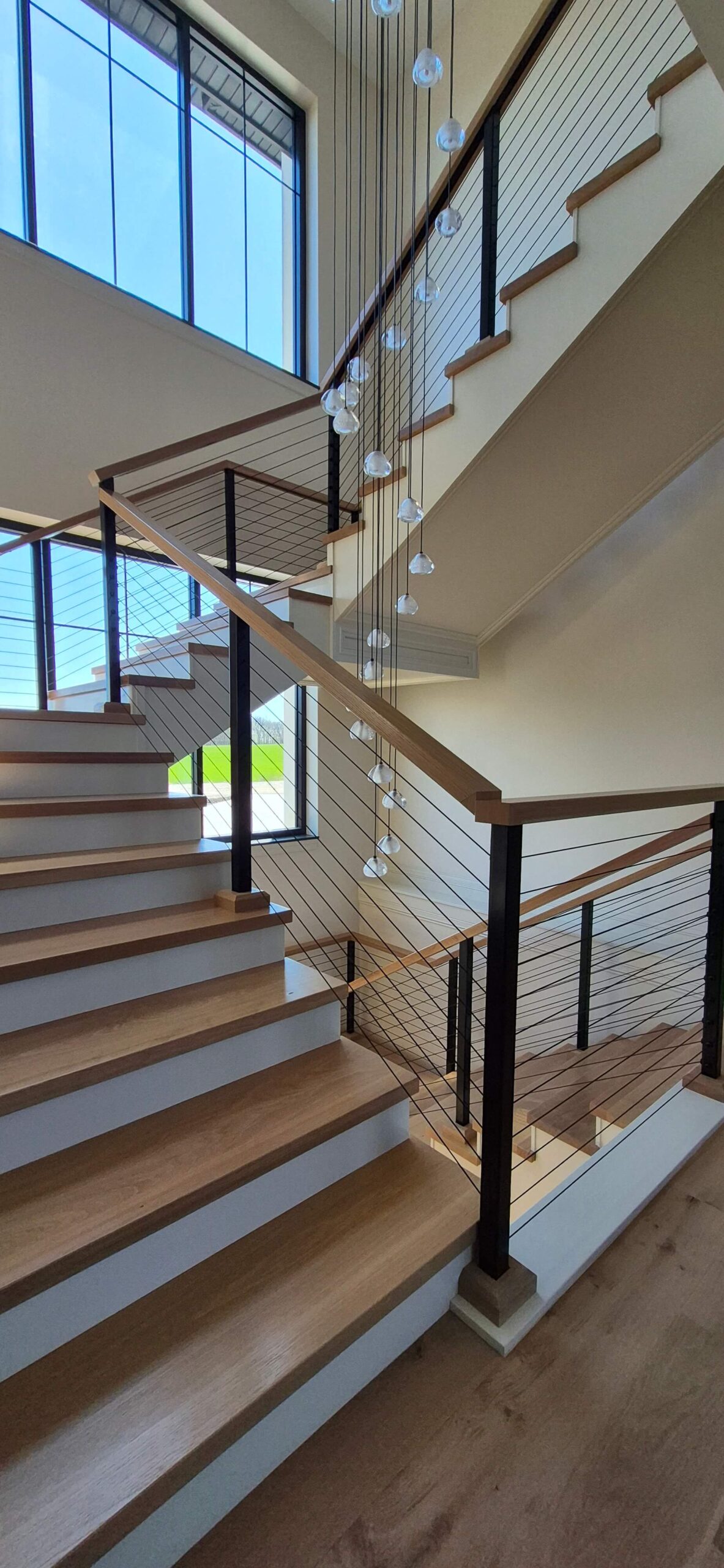 Creative Cabinetry Bloomfield Iowa A modern wooden staircase with cable railing and hanging pendant lights, located in a bright interior with large windows.
